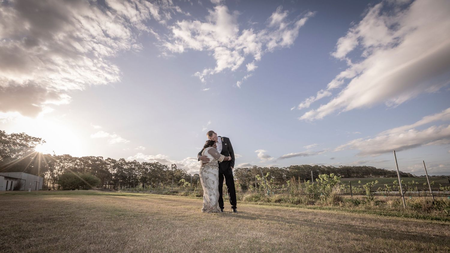 A couple shares a romantic moment at sunset on a hilltop overlooking scenic vineyards and countryside.