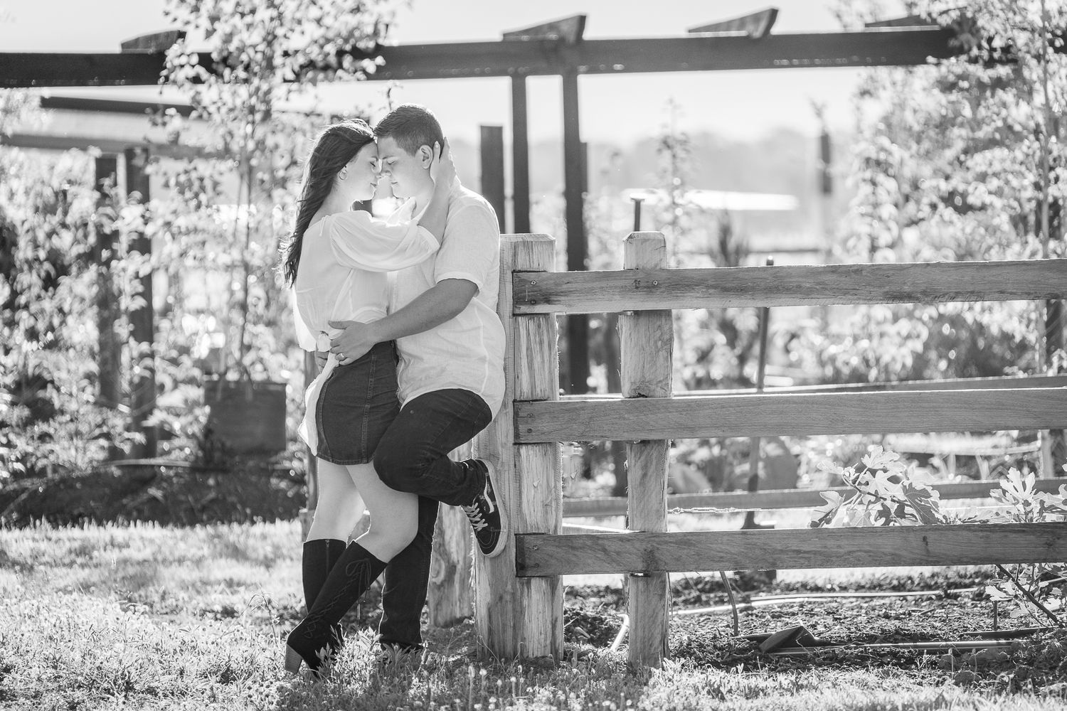 A couple embraces intimately on a wooden bench in a sunlit outdoor setting, captured in black and white.