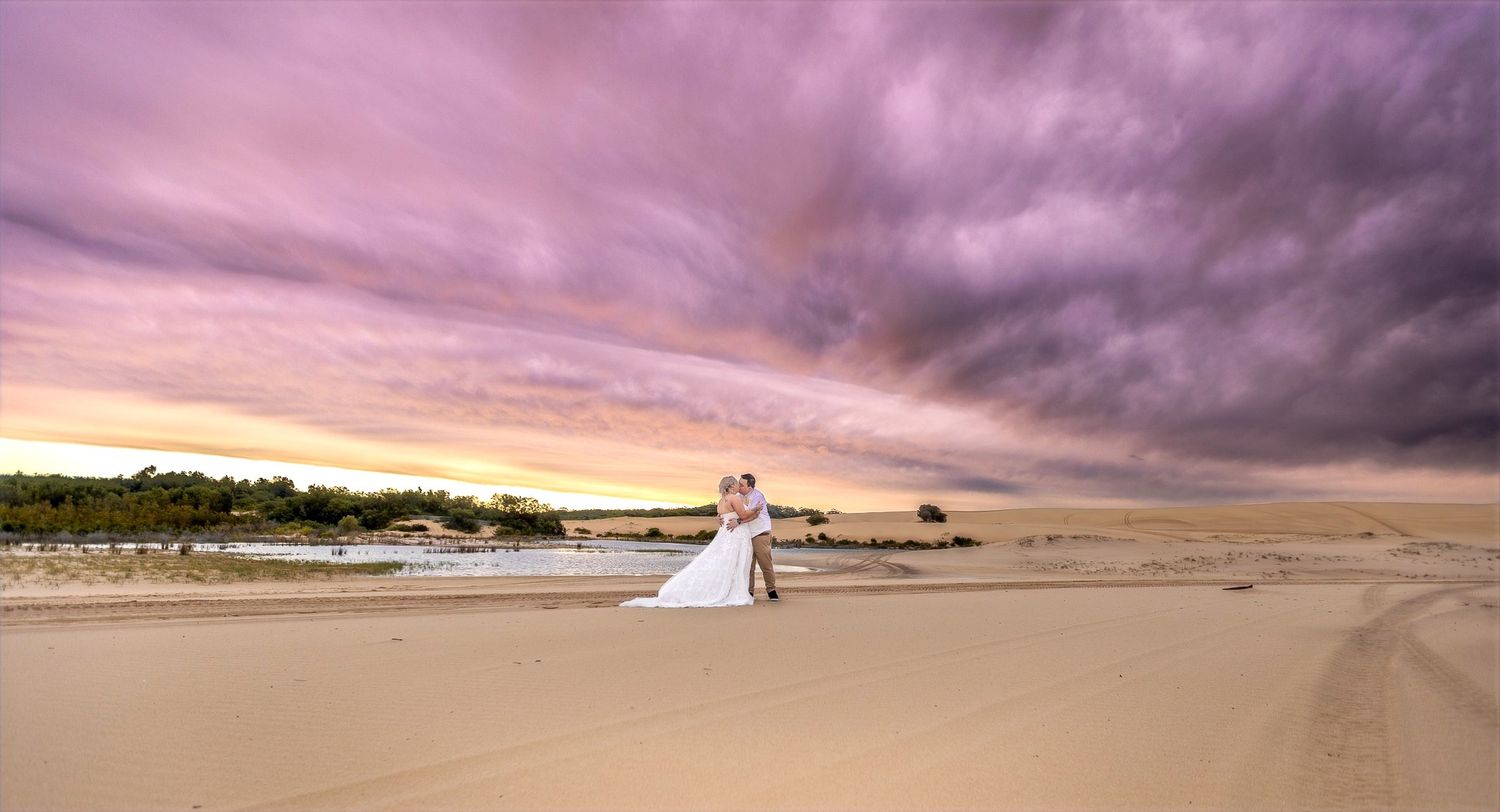 A couple in wedding attire stands on a sandy beach during dramatic sunset with purple and orange clouds.