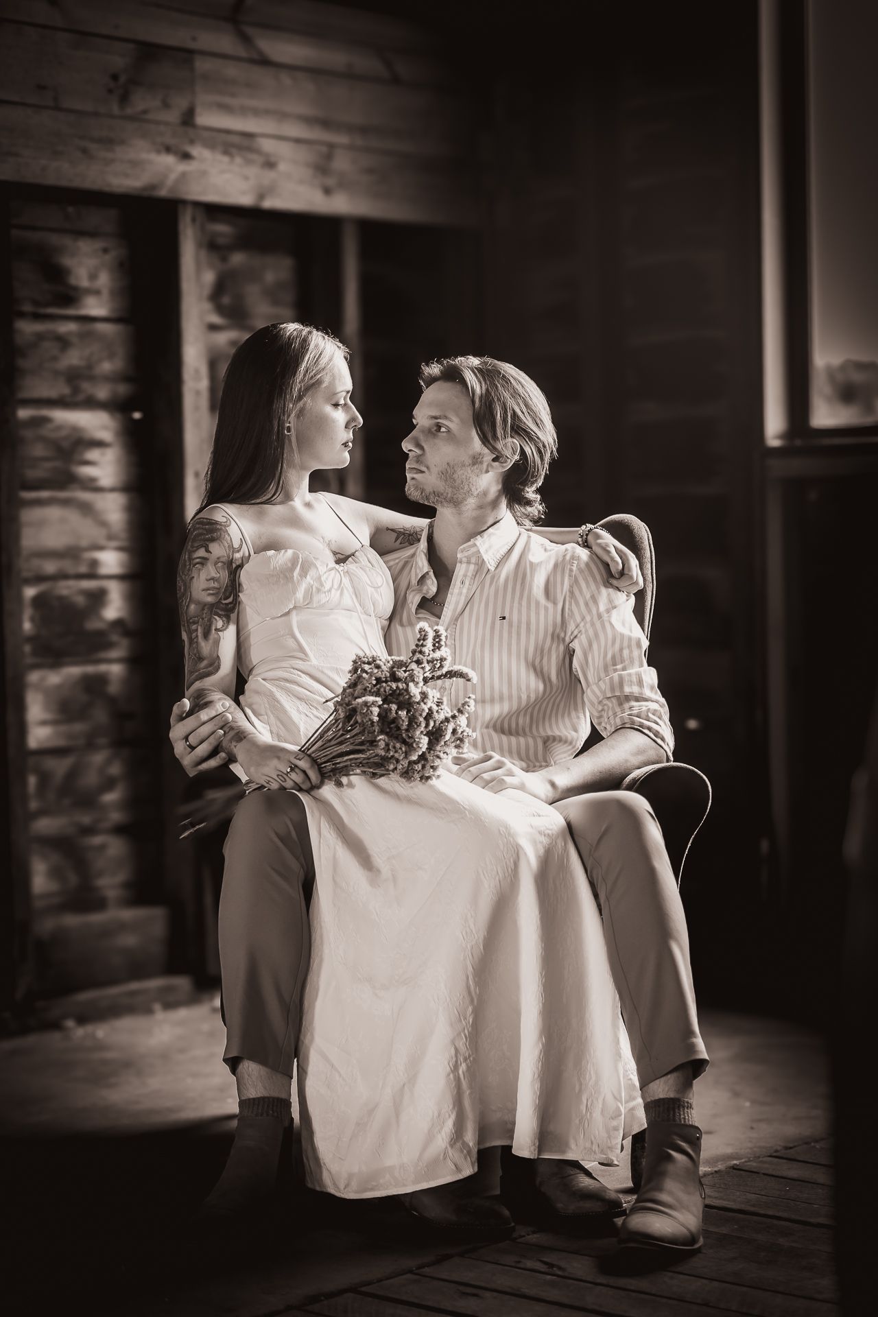 Sepia-toned portrait of a couple sitting together, woman in white dress with bouquet, man in striped shirt, rustic wooden interior.