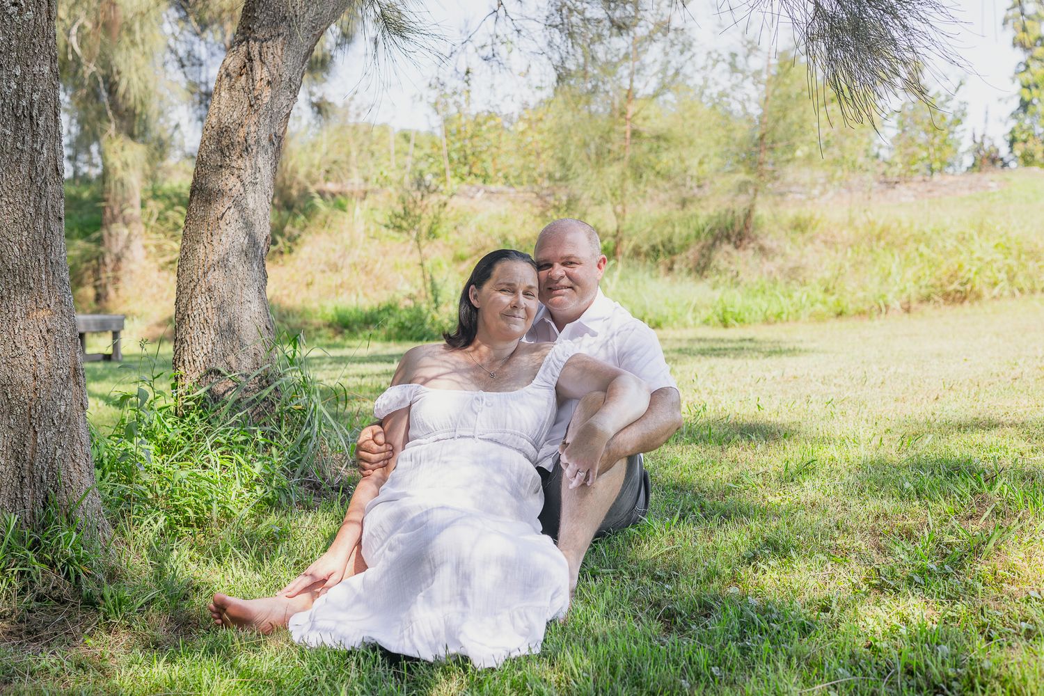 Couple sitting together under a tree on green grass, woman in white dress, man in white shirt, outdoor maternity photo.