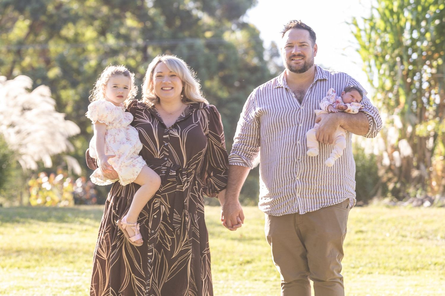 Happy family with two young children walking outdoors in a sunny park, parents smiling and holding babies.