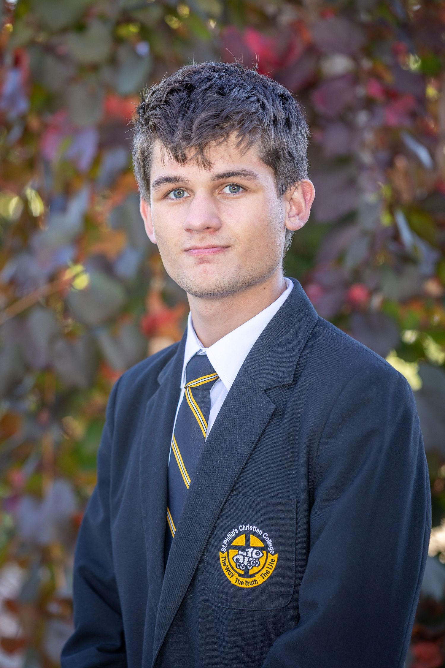 Young male student in navy school blazer with crest and striped tie poses outdoors against autumn foliage backdrop.