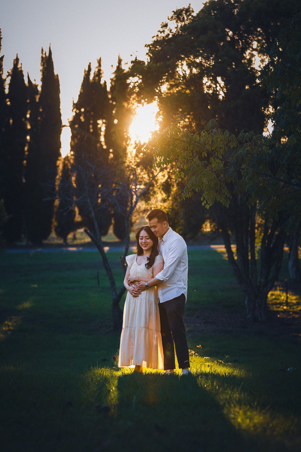 Couple embracing at golden hour sunset in lush green park, woman in blush dress, man in white shirt.