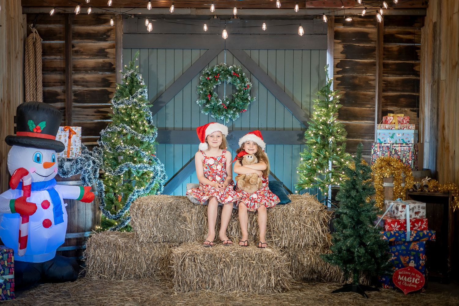 A rustic barn decorated for Christmas with hay bales, lit trees, an inflatable snowman, and festive holiday decor.