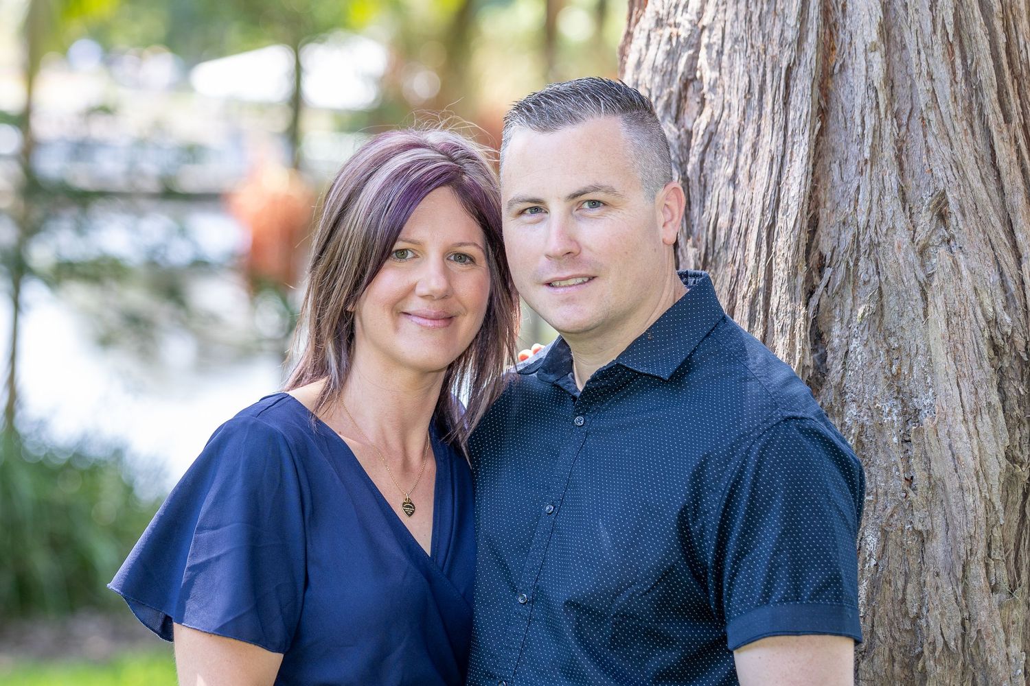 A couple smiles while standing next to a large tree trunk outdoors in a park-like setting by water.
