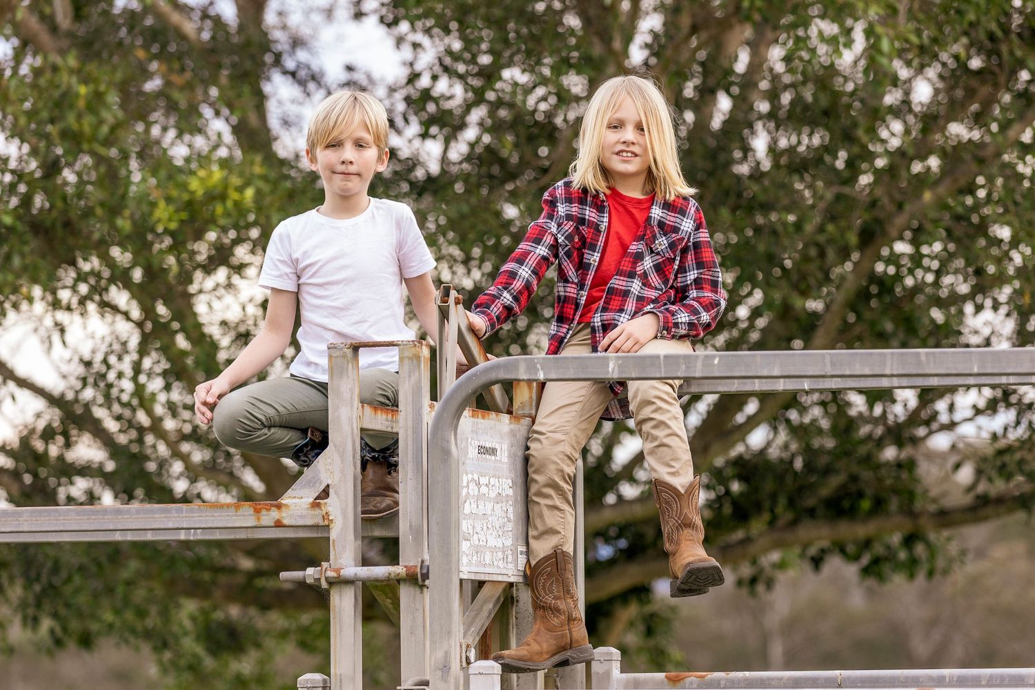 Two children sit playfully on metal playground equipment in an outdoor setting.