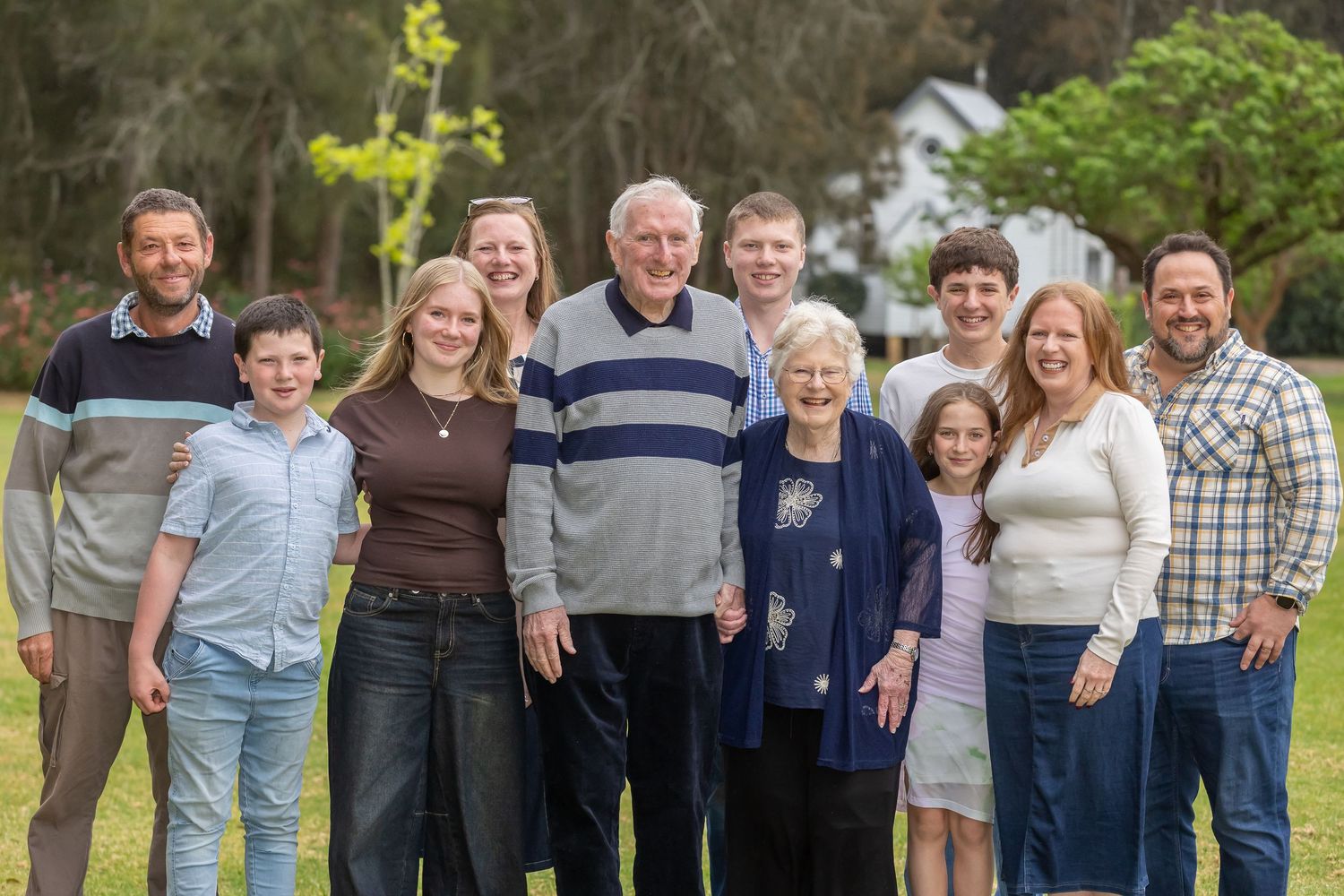 Large multigenerational family group smiling together outdoors on green lawn with trees in background.
