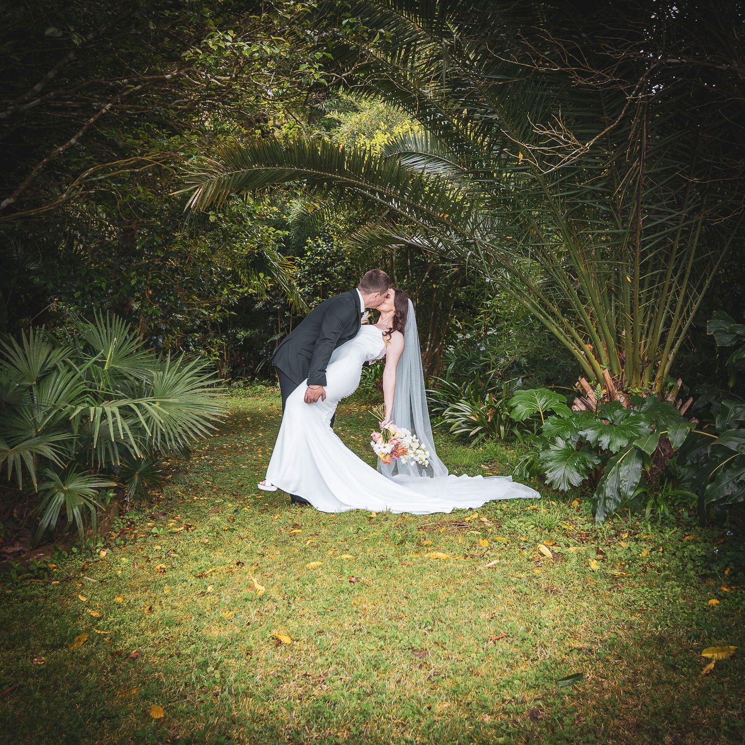 A romantic moment under palm trees in a lush garden setting with scattered autumn leaves on green grass.