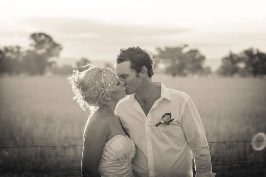 A romantic moment captured in black and white as a couple embraces in a misty field with wooden fence in background.