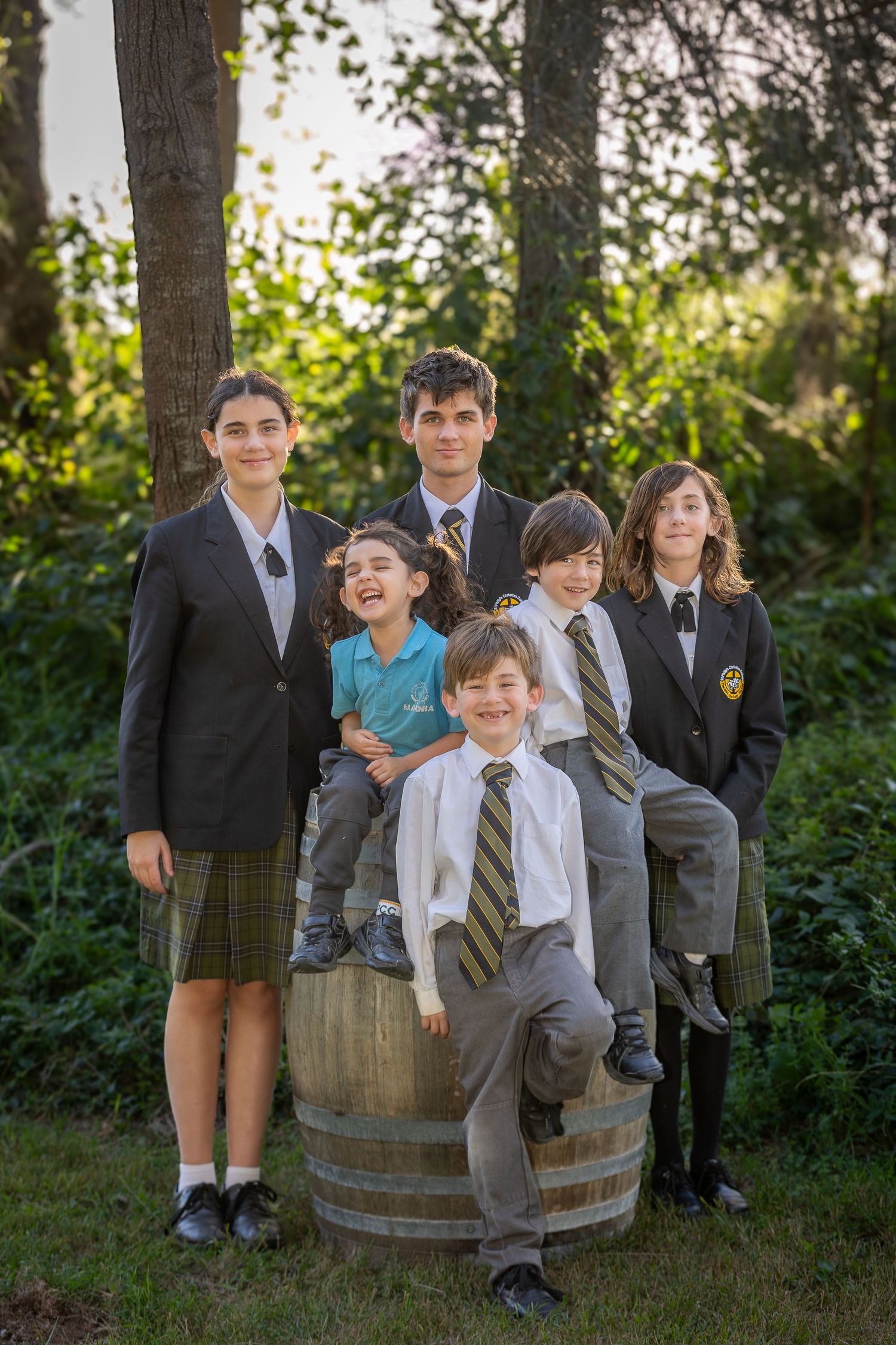 Group of seven children in school uniforms posing outdoors around a wooden barrel among trees.