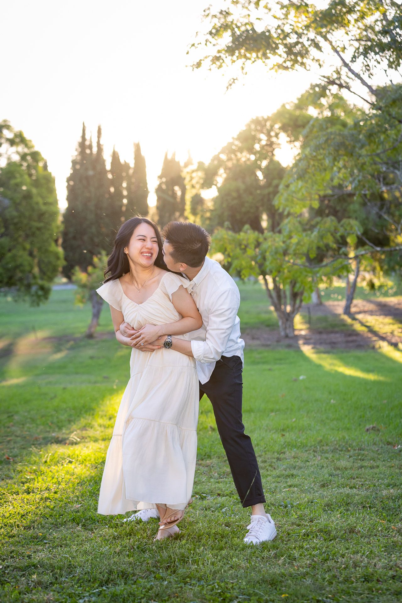 Couple sharing a romantic moment in a sunlit park, woman in white dress laughing as man embraces her from behind.