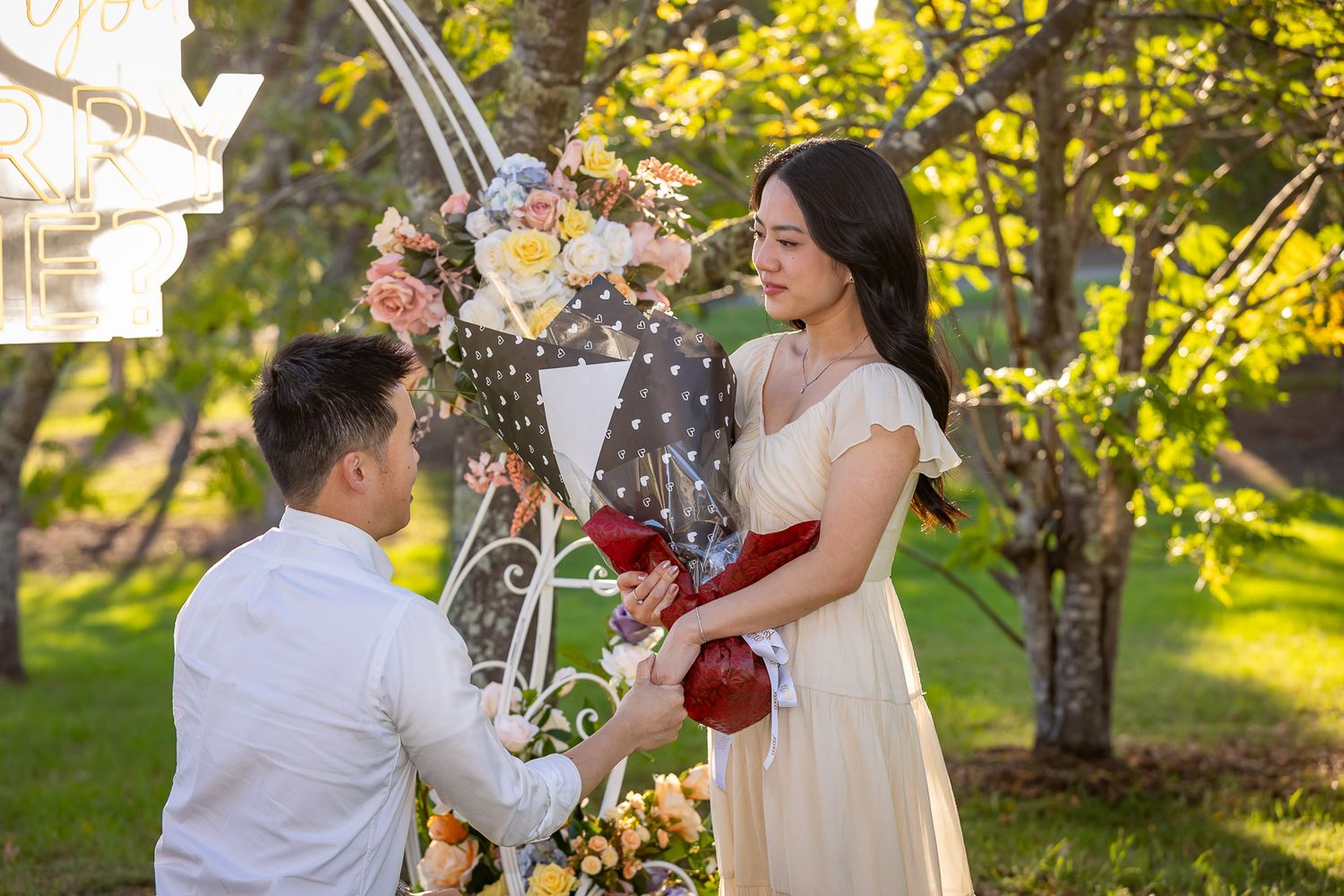 A man proposes to a woman in a garden, offering red roses near a floral arch on a sunny day.
