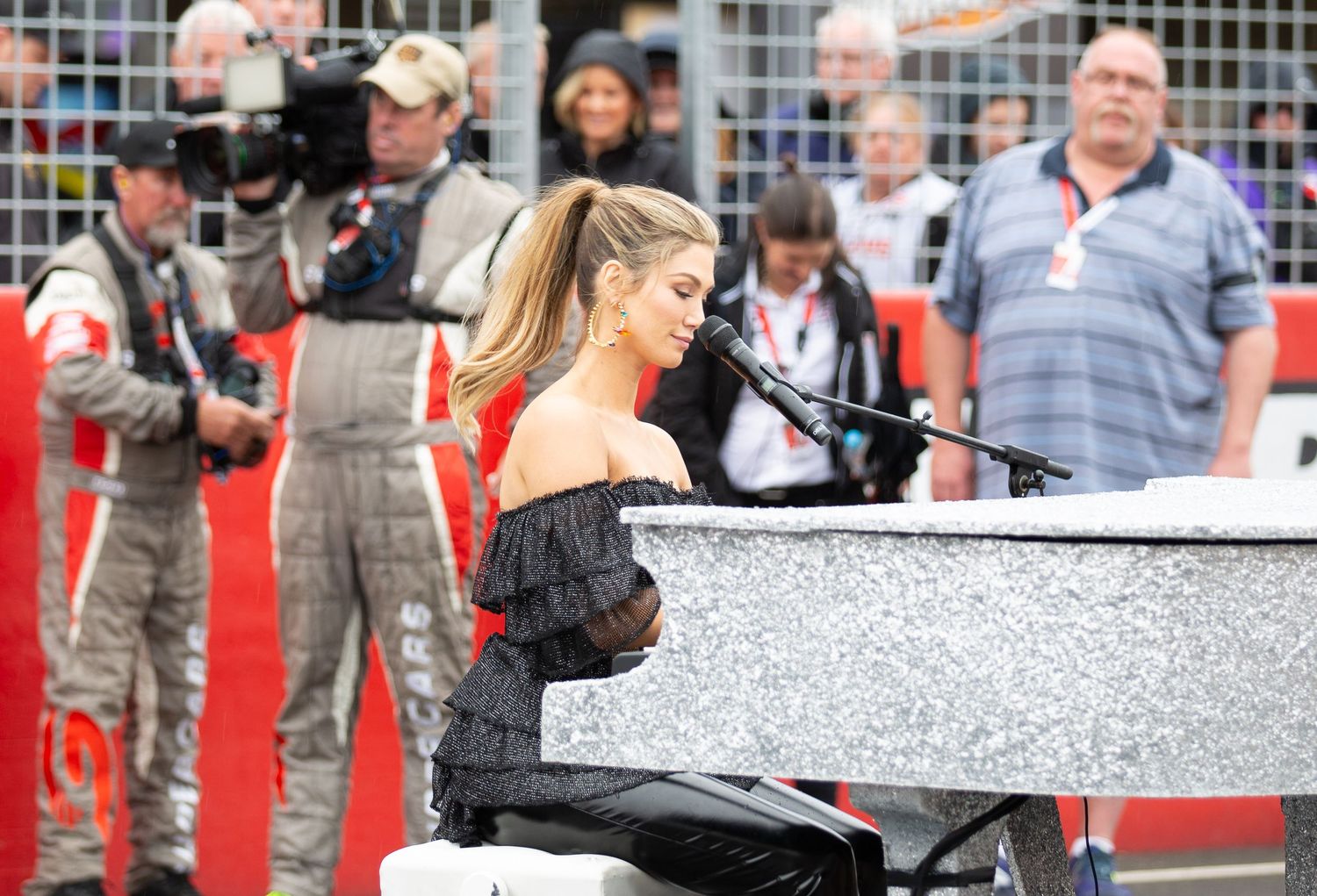 Delta Goodrem in a black and red outfit plays a glittering white grand piano during an outdoor event with cameras rolling.