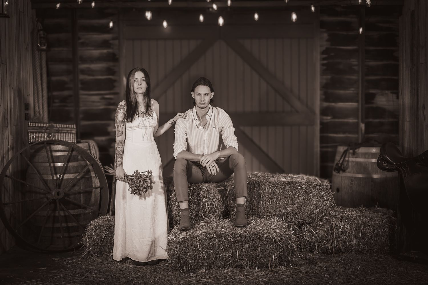 Sepia-toned photo of a bride and groom posing in a rustic barn with hay bales and string lights.