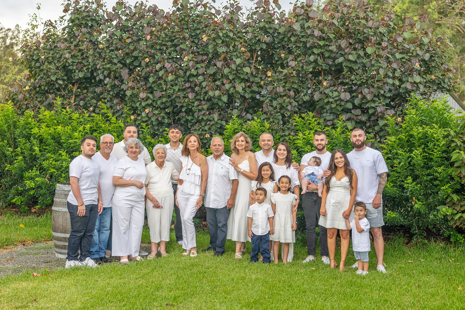 Large family group portrait outdoors in white outfits, posing together on green lawn with lush garden background.