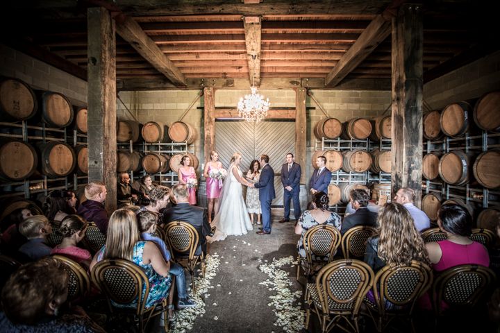 A romantic wedding ceremony takes place in a rustic winery barrel room with flower petals scattered on the ground.