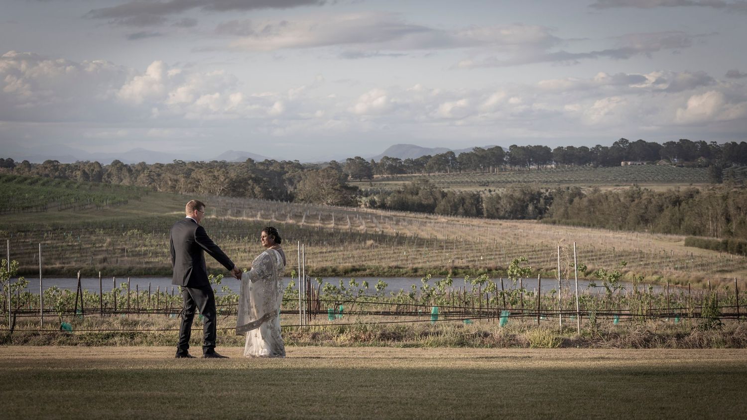 Couple holds hands while admiring scenic vineyard landscape with rolling hills and cloudy sky in countryside.