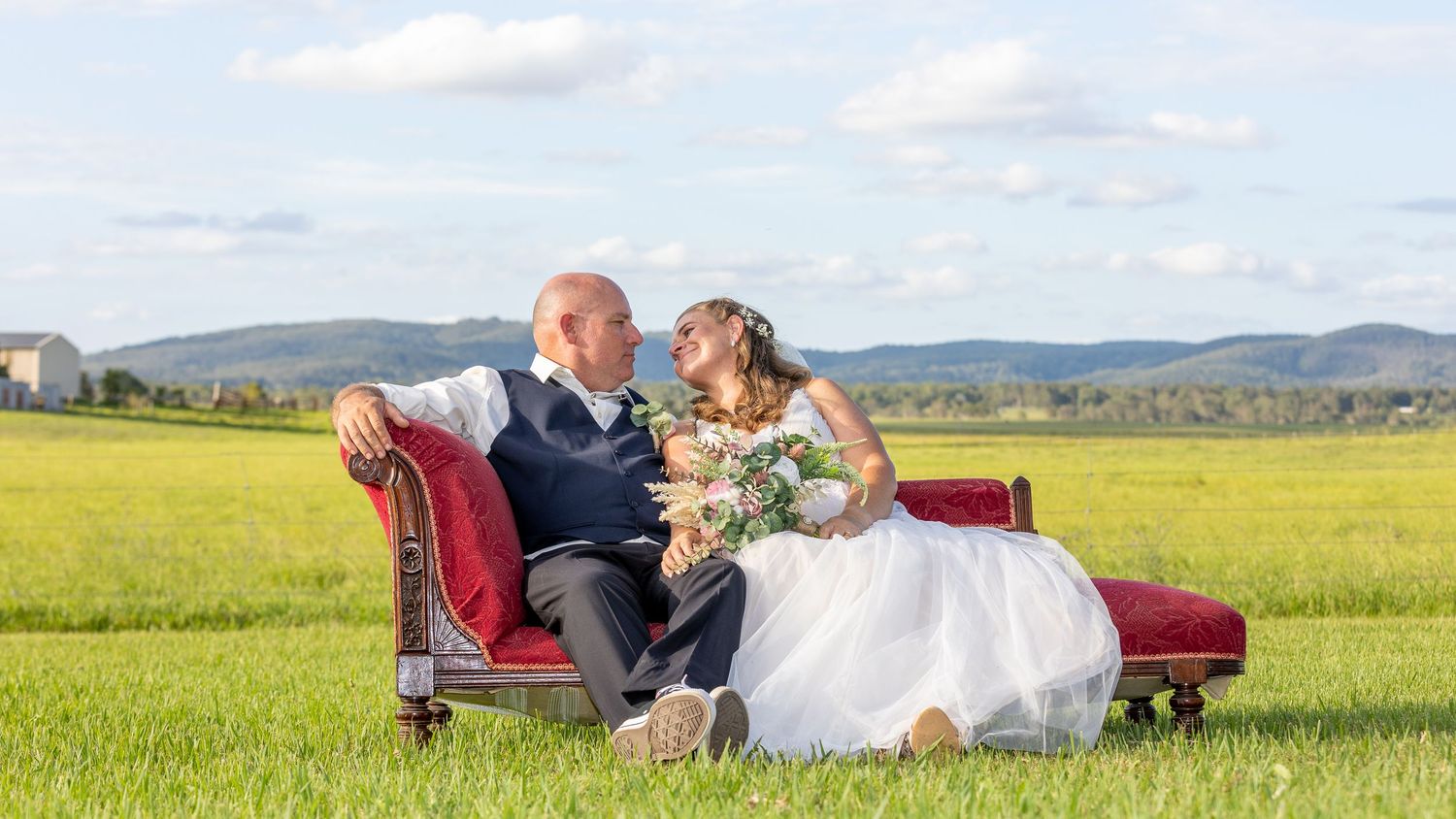 Wedding couple rests in a peaceful meadow with mountain views and a rustic burgundy sofa during a sunset photo session.