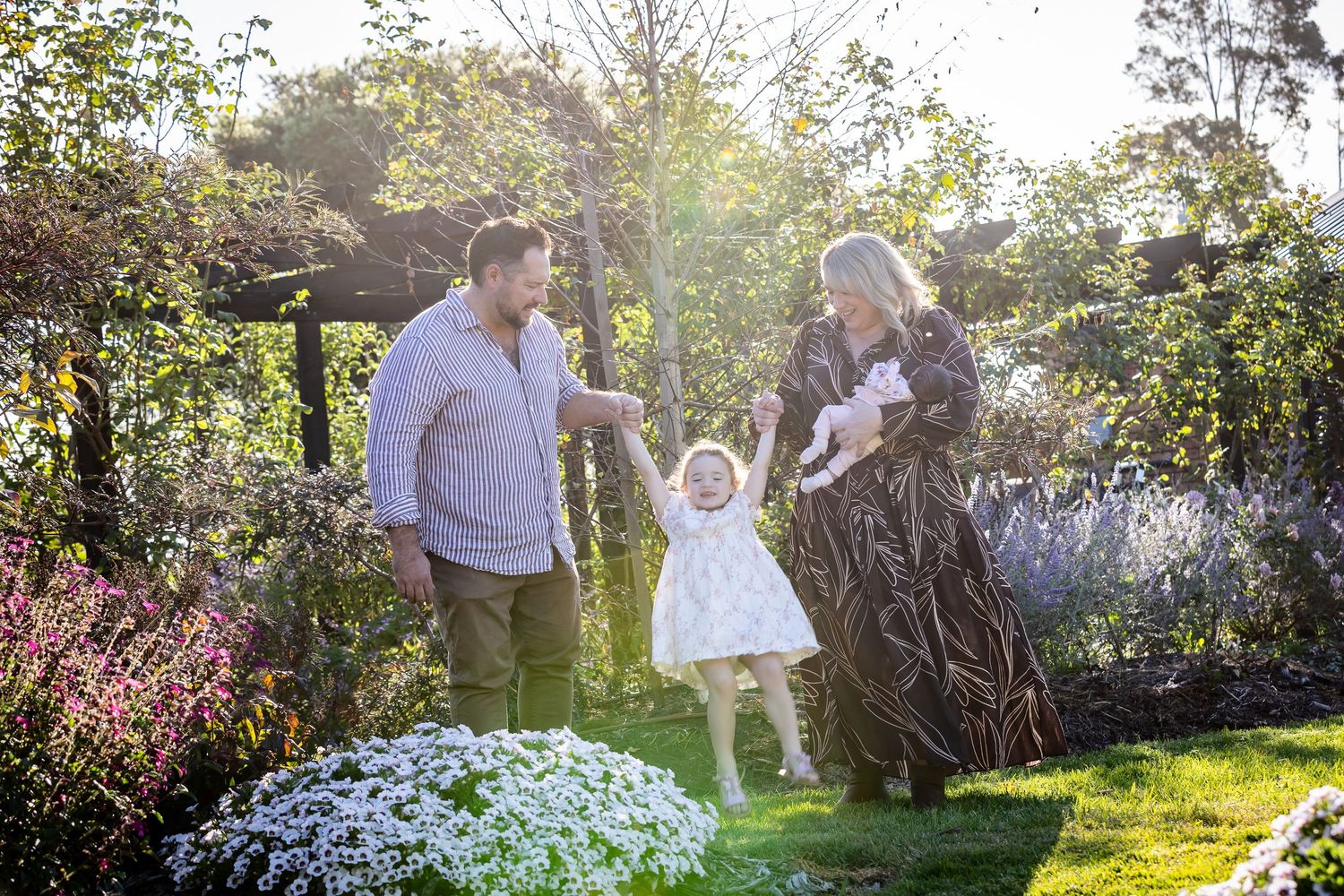 Family with toddler and newborn enjoying a sunny garden, parents swinging young child in white dress.