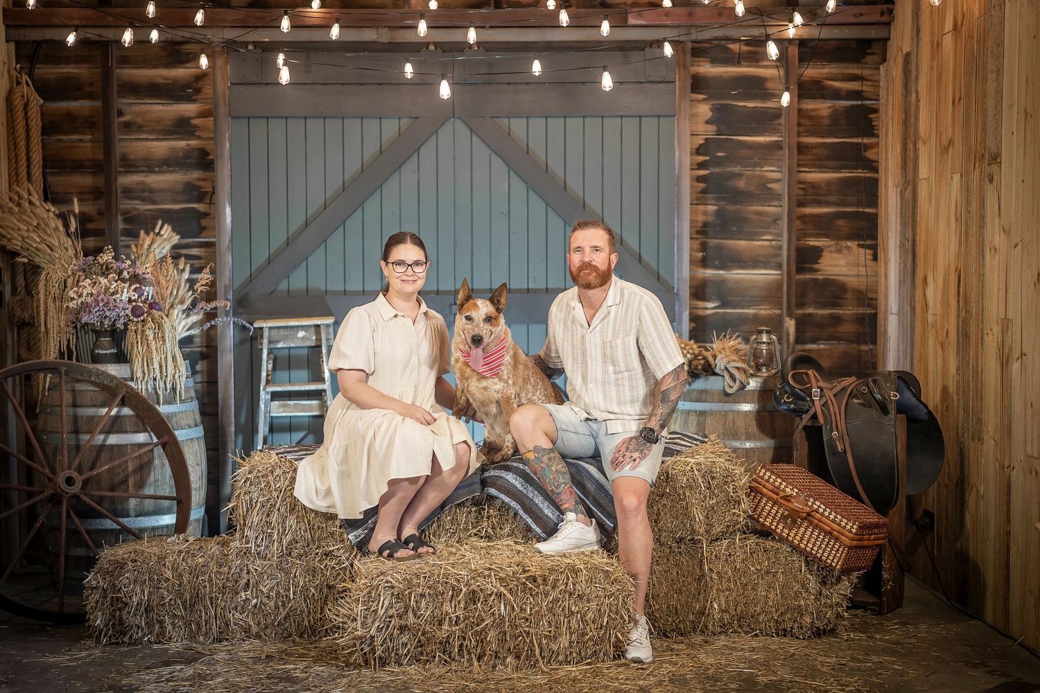Family with dog posing on hay bales in rustic barn setting with string lights and vintage decor.
