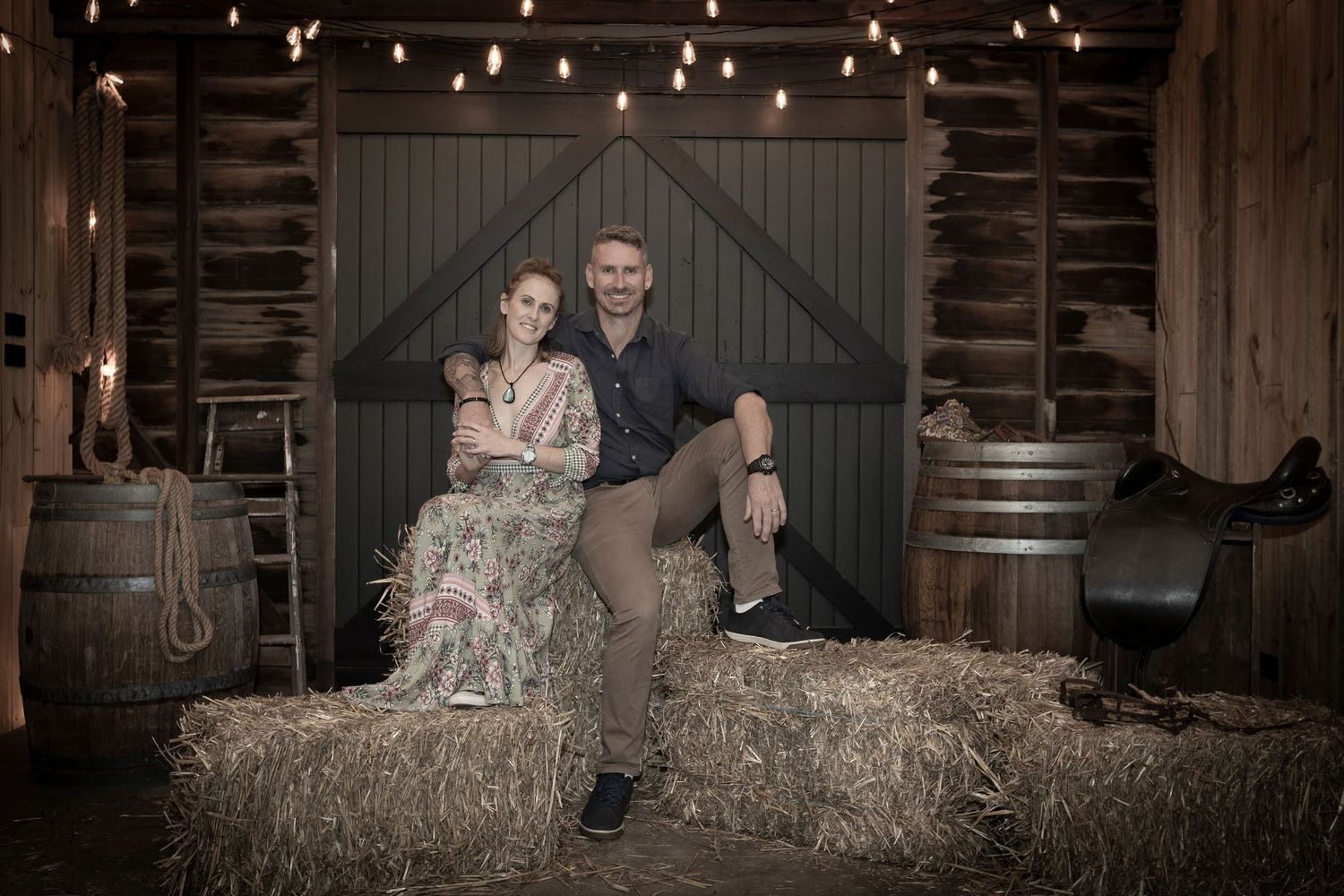 Couple posing on hay bales in a rustic barn with wooden backdrop and string lights.