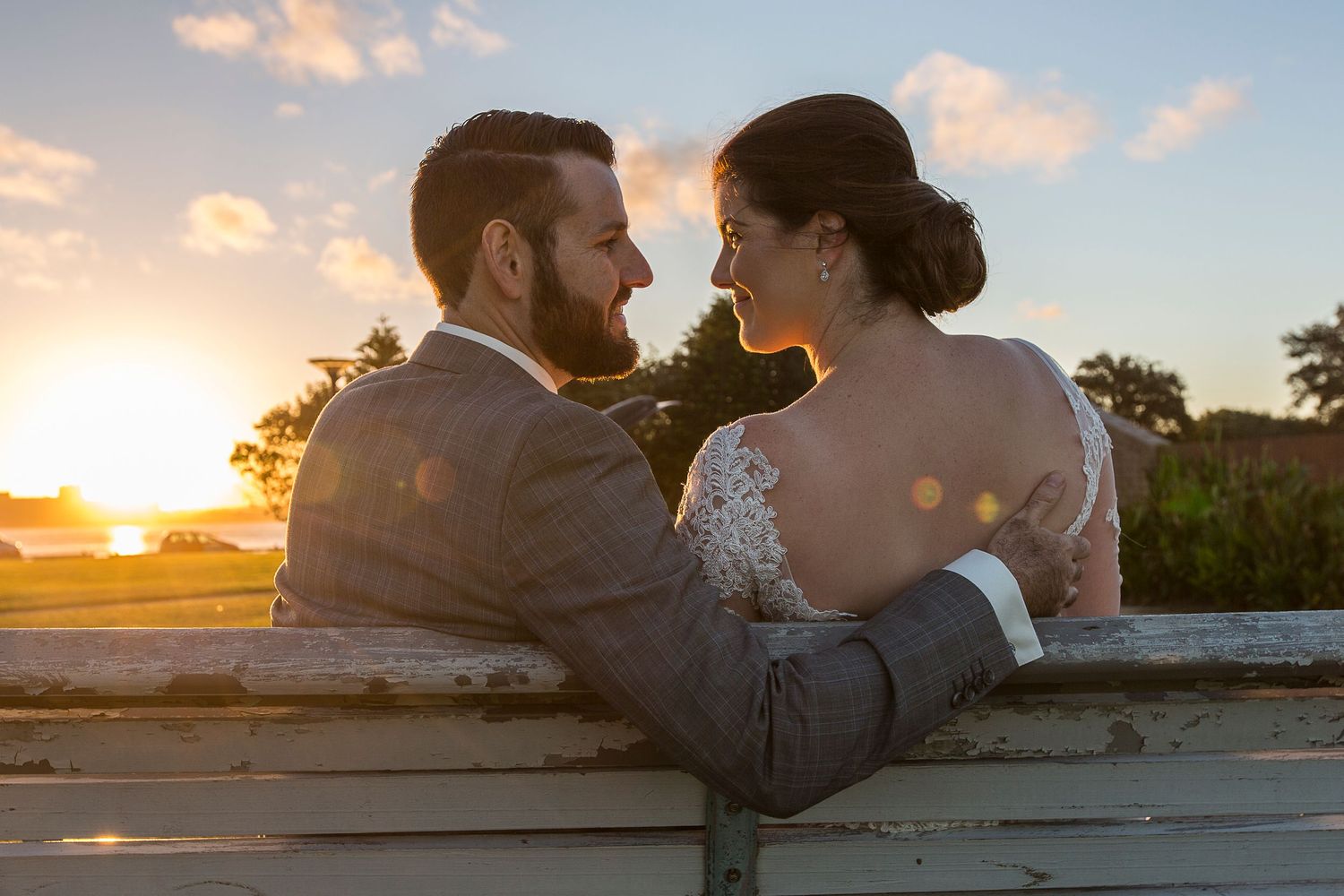 A romantic couple embraces while sitting together at sunset, silhouetted against a warm golden sky and palm trees.