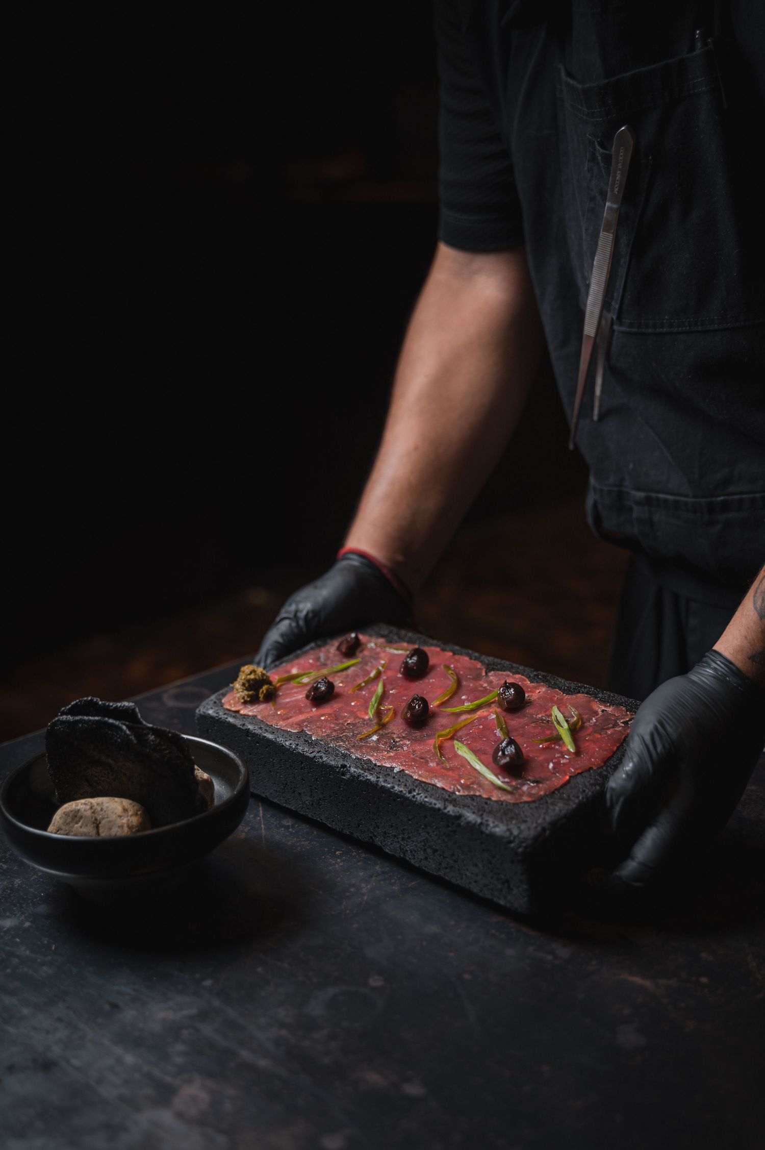 Dark moody food photography sequence showing hands preparing raw beef tartare with garnishes on black slate surface.