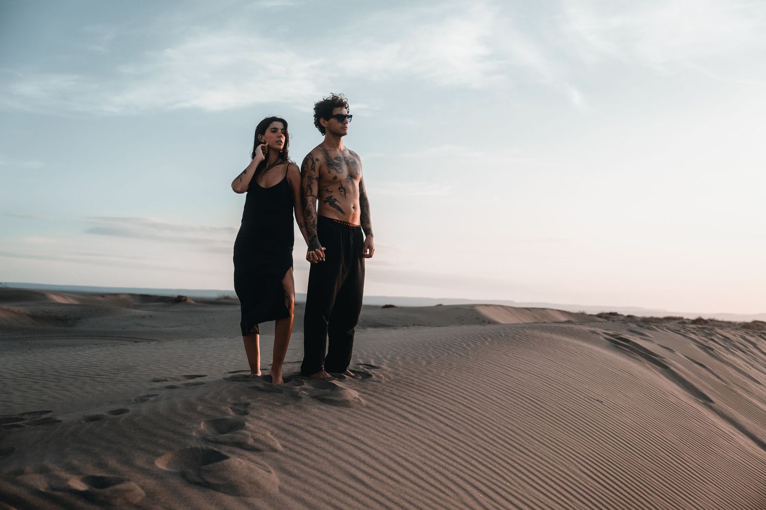A couple stands together on sand dunes during sunset in a desert landscape.