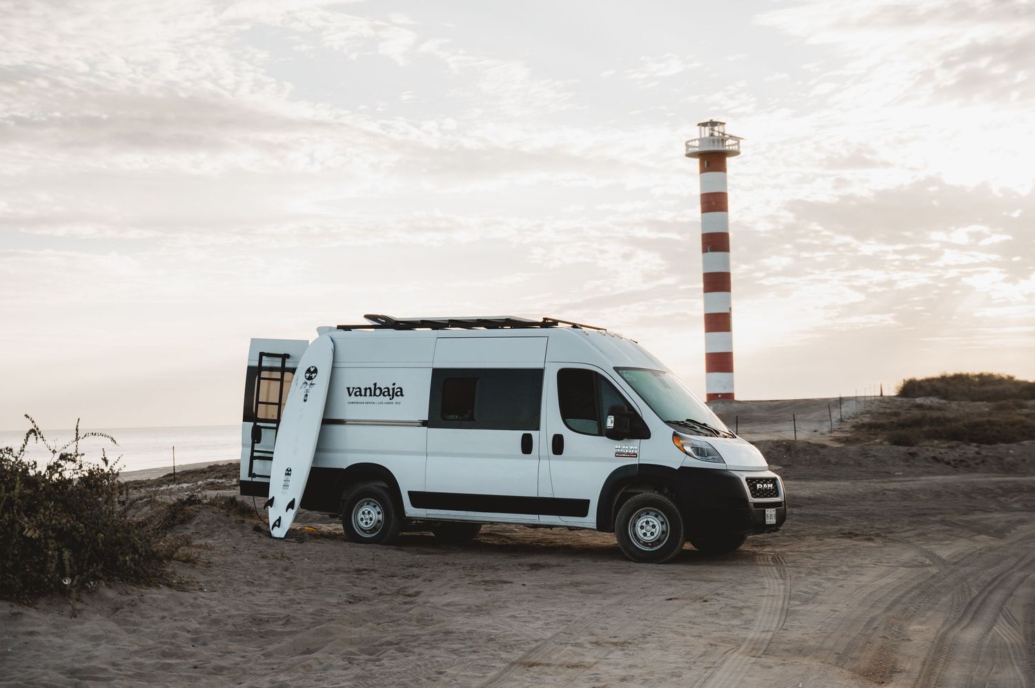 White camper van parked near striped lighthouse against cloudy coastal sky.