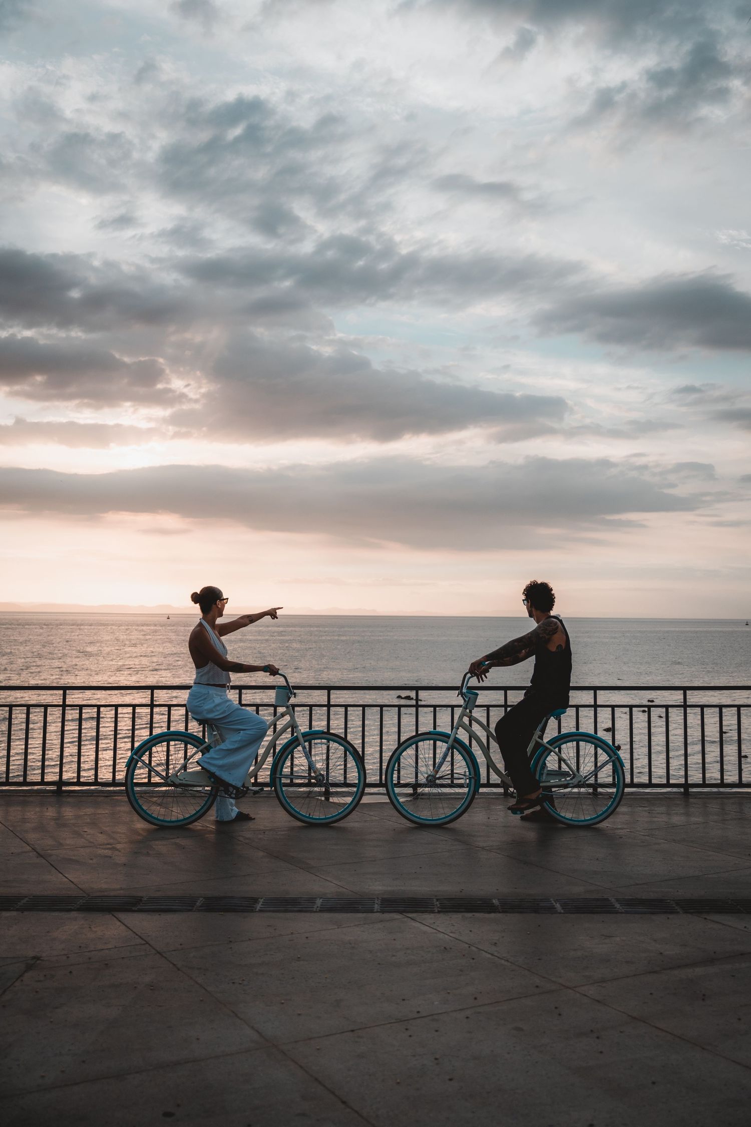 Two cyclists stand with their bikes on a seaside promenade at sunset.