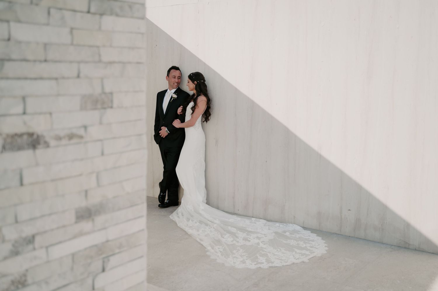 Elegant wedding couple poses against modern white stone wall architecture.