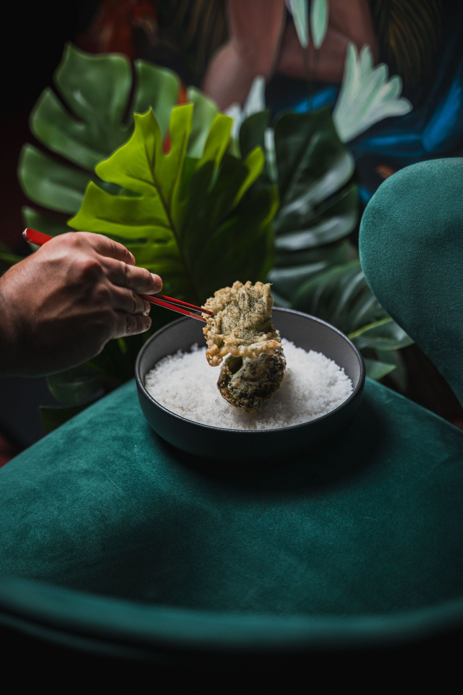 Hand holding red chopsticks serving food from bowl filled with salt or ice against dark background.