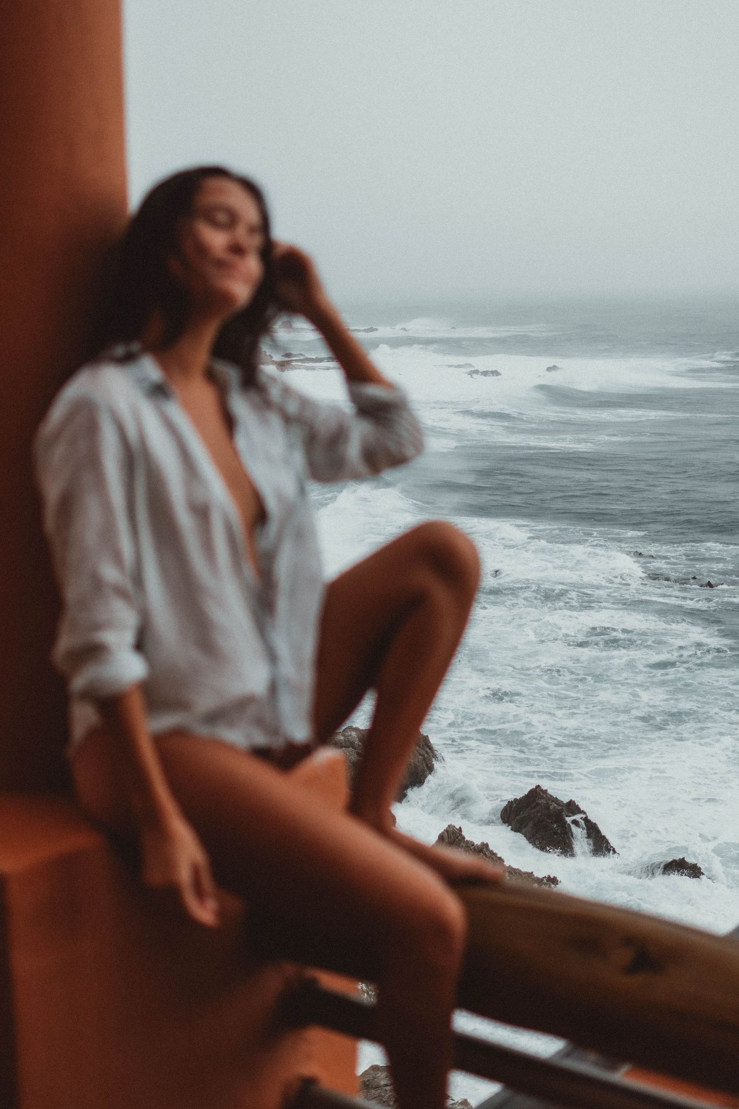 A moody coastal photoshoot featuring a white button-down shirt against dramatic ocean waves and rocks.