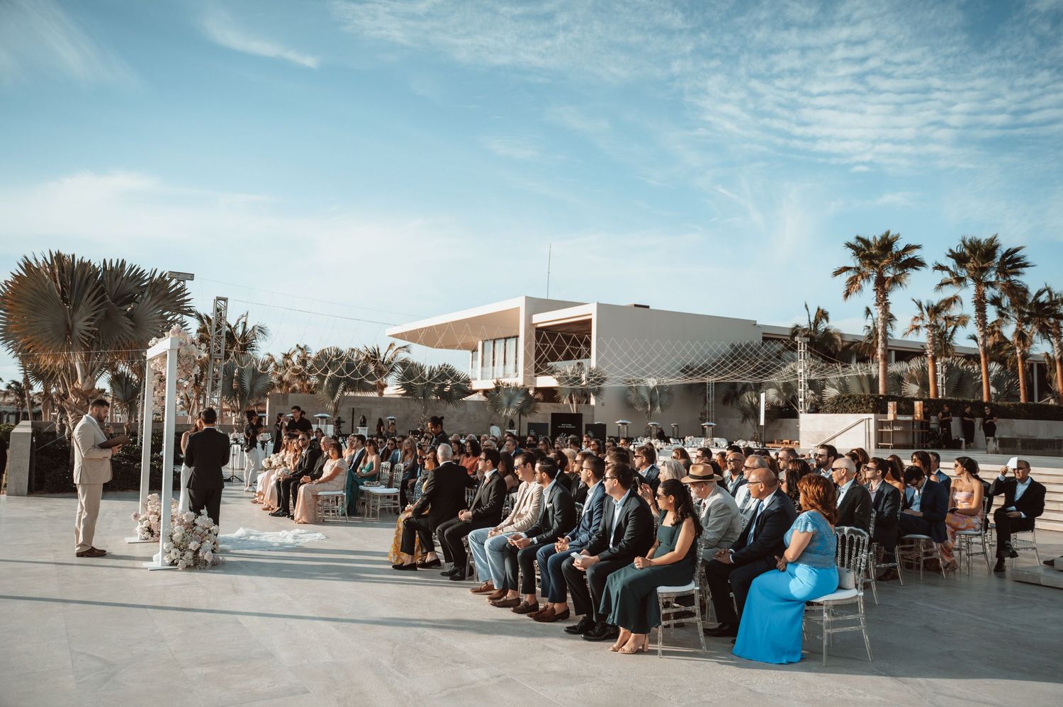 Outdoor wedding ceremony setup with palm trees and rows of white chairs under bright blue sky.
