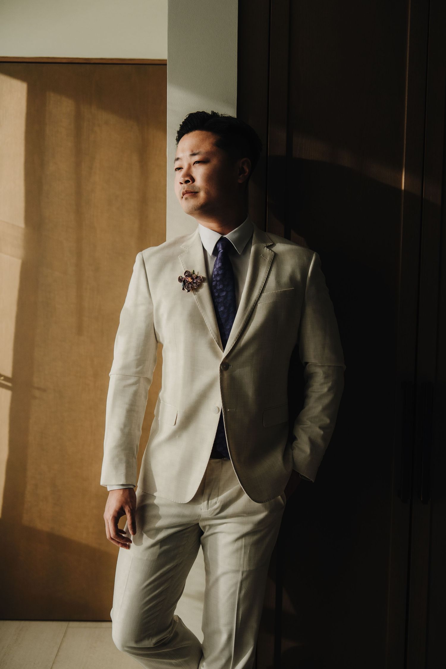 Groom in light-colored suit stands in dramatic lighting against neutral wall.