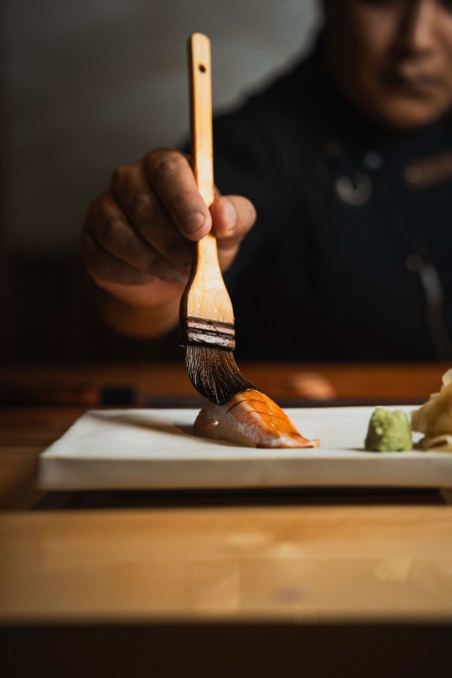 Chef applying sauce with brush to sushi piece on white plate.
