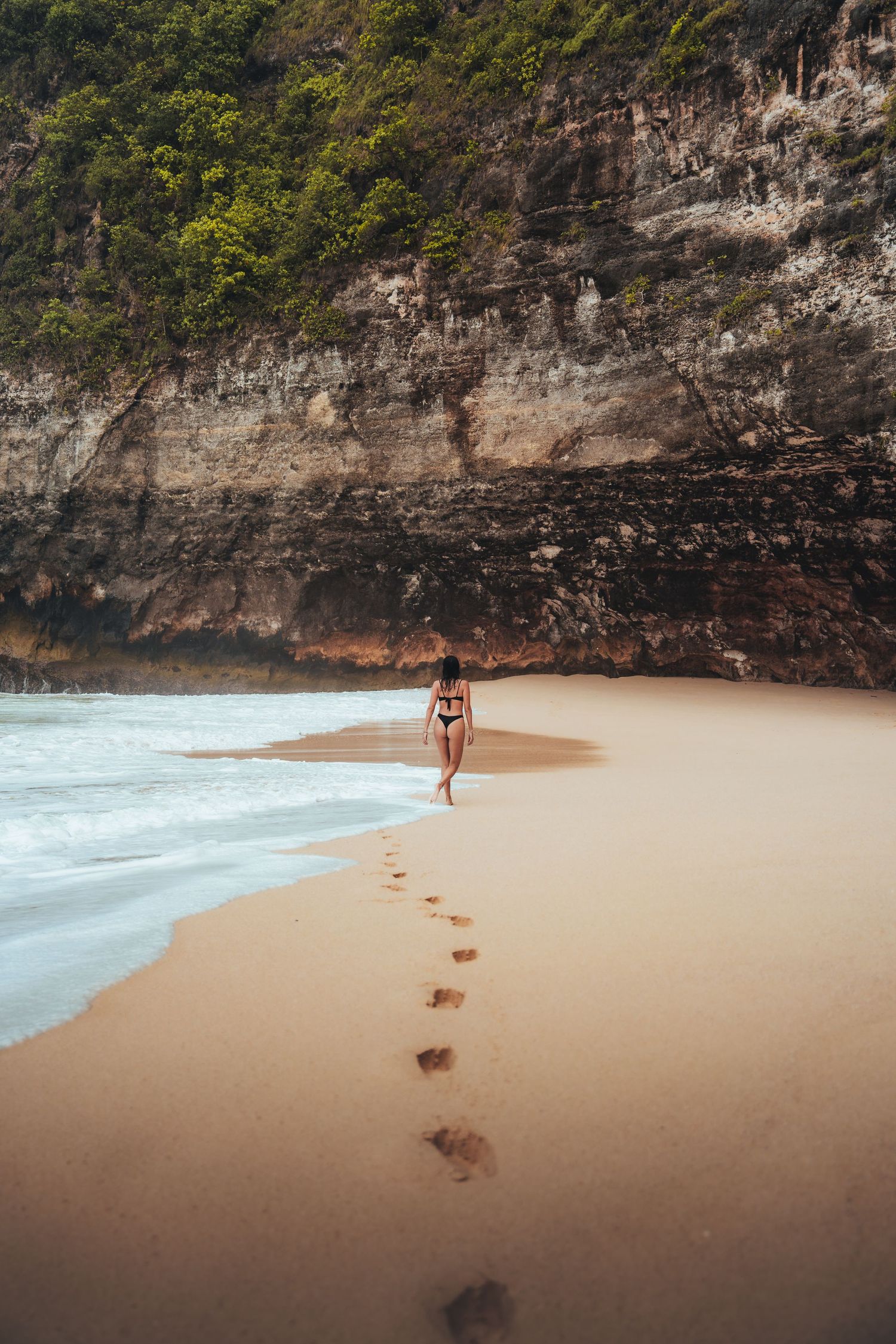 Footprints trailing across a sandy beach with rocky cliffs and ocean waves.