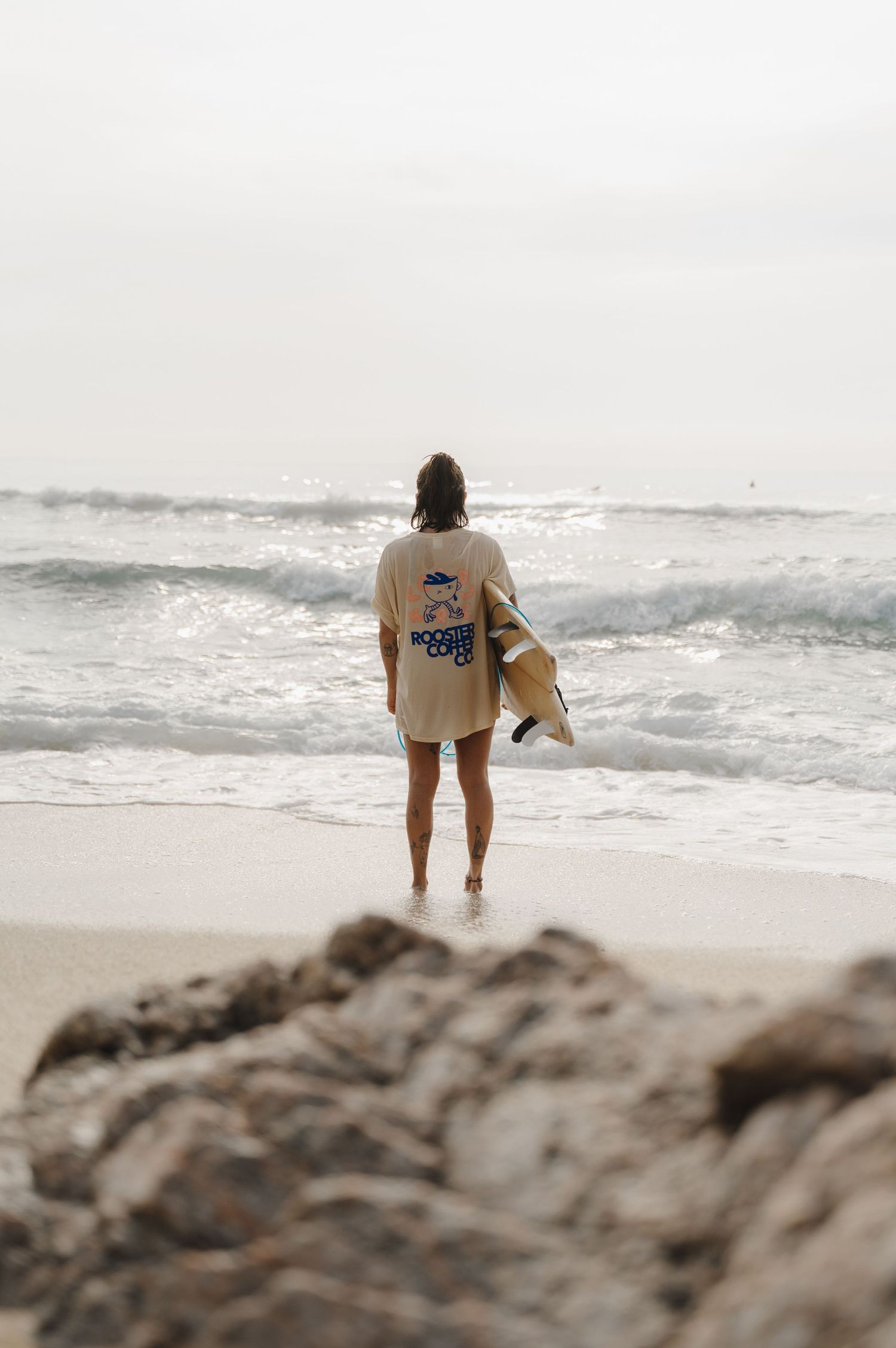 Person carrying surfboard walks along misty beach shoreline.