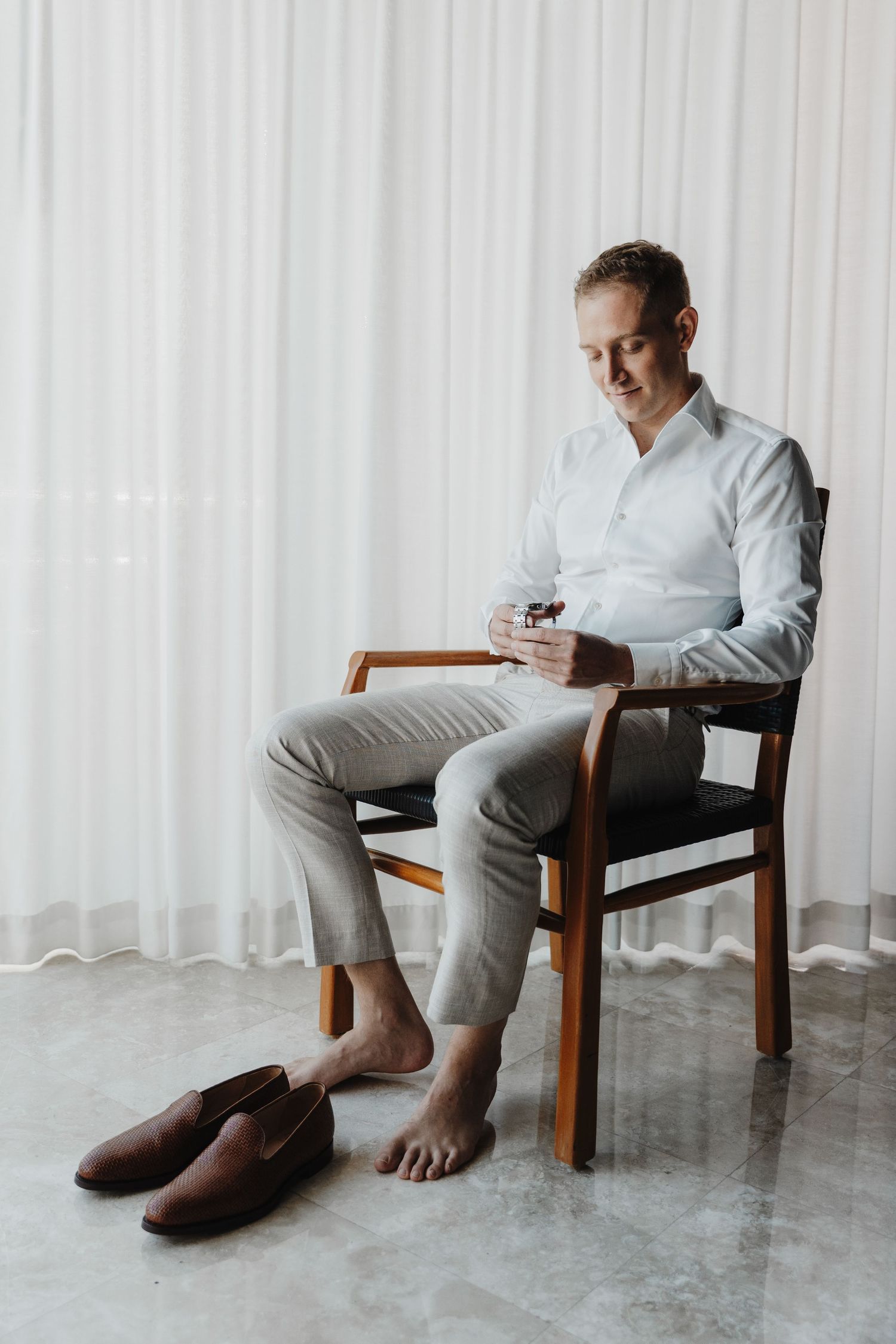 Person in grey suit and white shirt sits barefoot in wooden chair against white curtain backdrop.