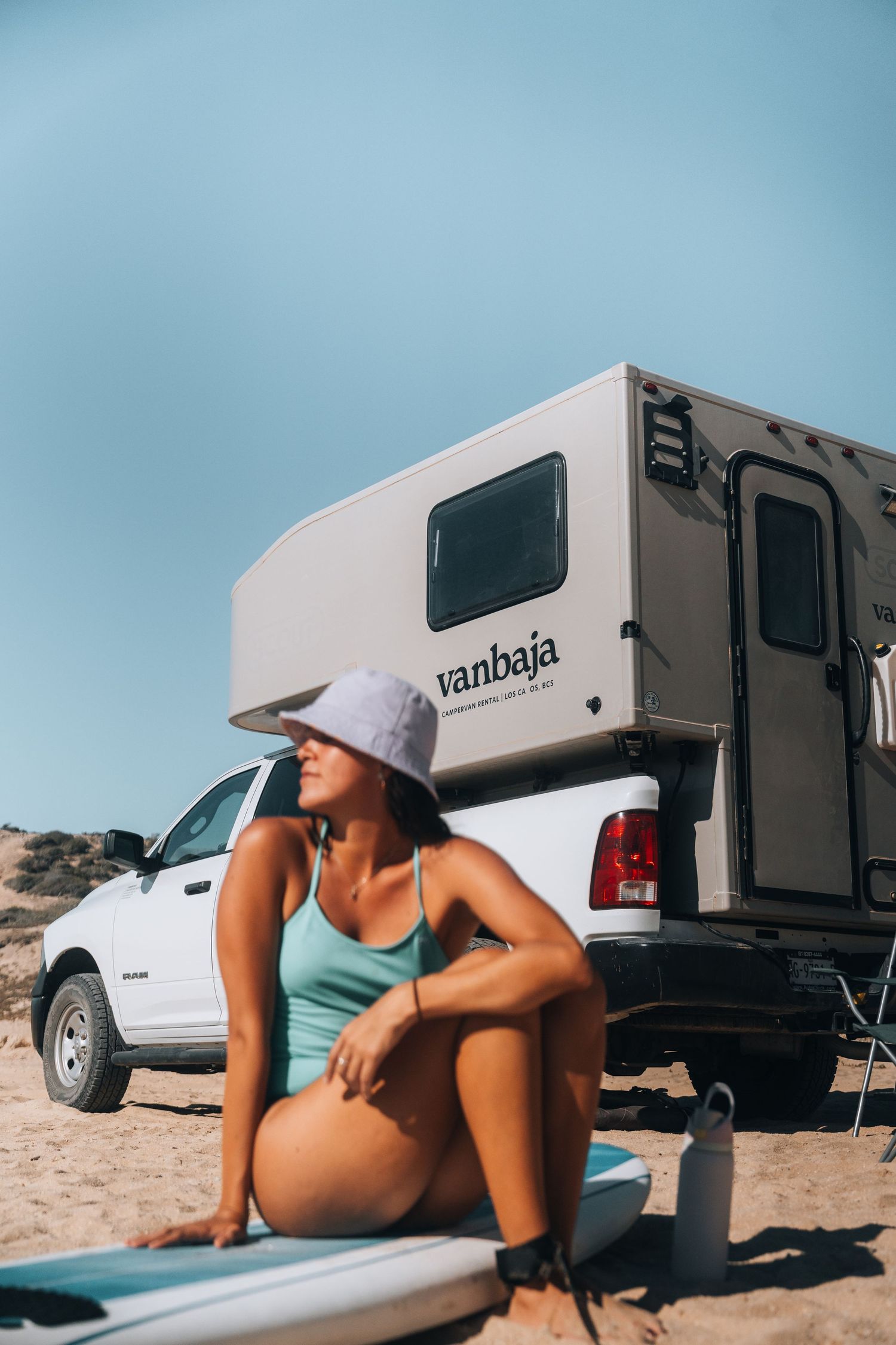 RV and truck parked on a sunny beach with someone lounging in swimwear.