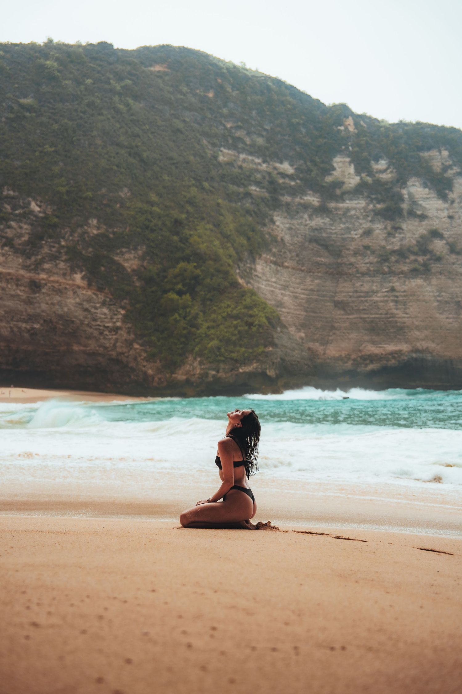 A person sits peacefully on a sandy beach with dramatic cliffs and turquoise waters in the background.