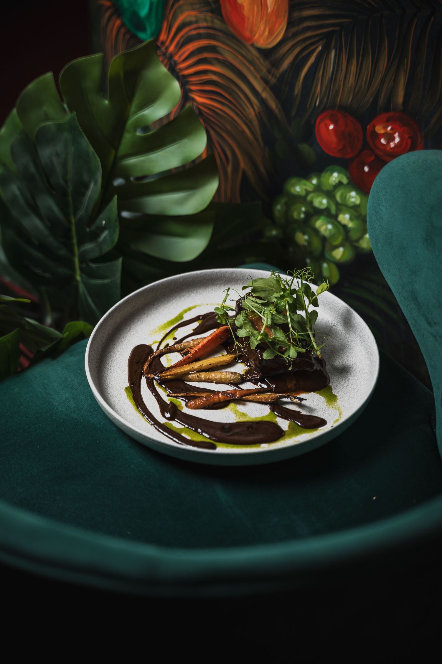 Elegantly plated dish with microgreens and dark sauce on white plate against tropical leaf backdrop.