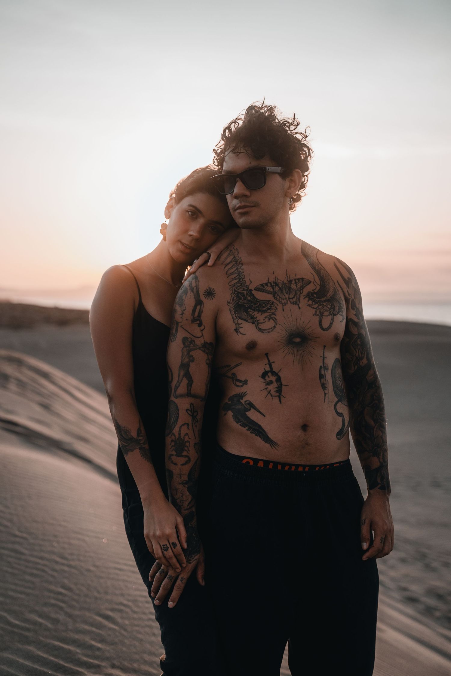 Two people embracing on sand dunes at sunset creating intimate silhouettes against the sky.