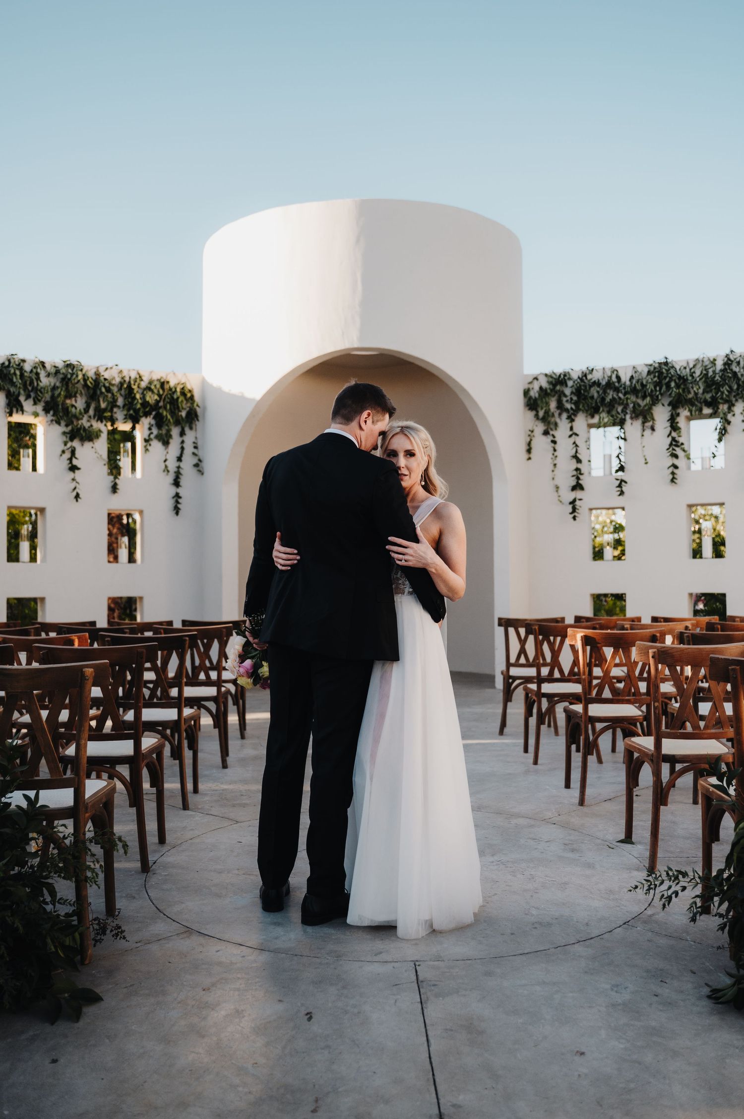 Wedding ceremony setup in a white arched venue with wooden chairs and floral decorations.
