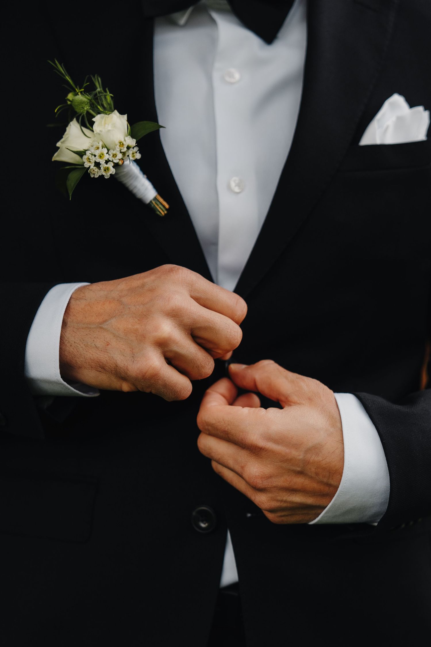 Groom in black tuxedo adjusts buttons with white boutonniere on lapel.
