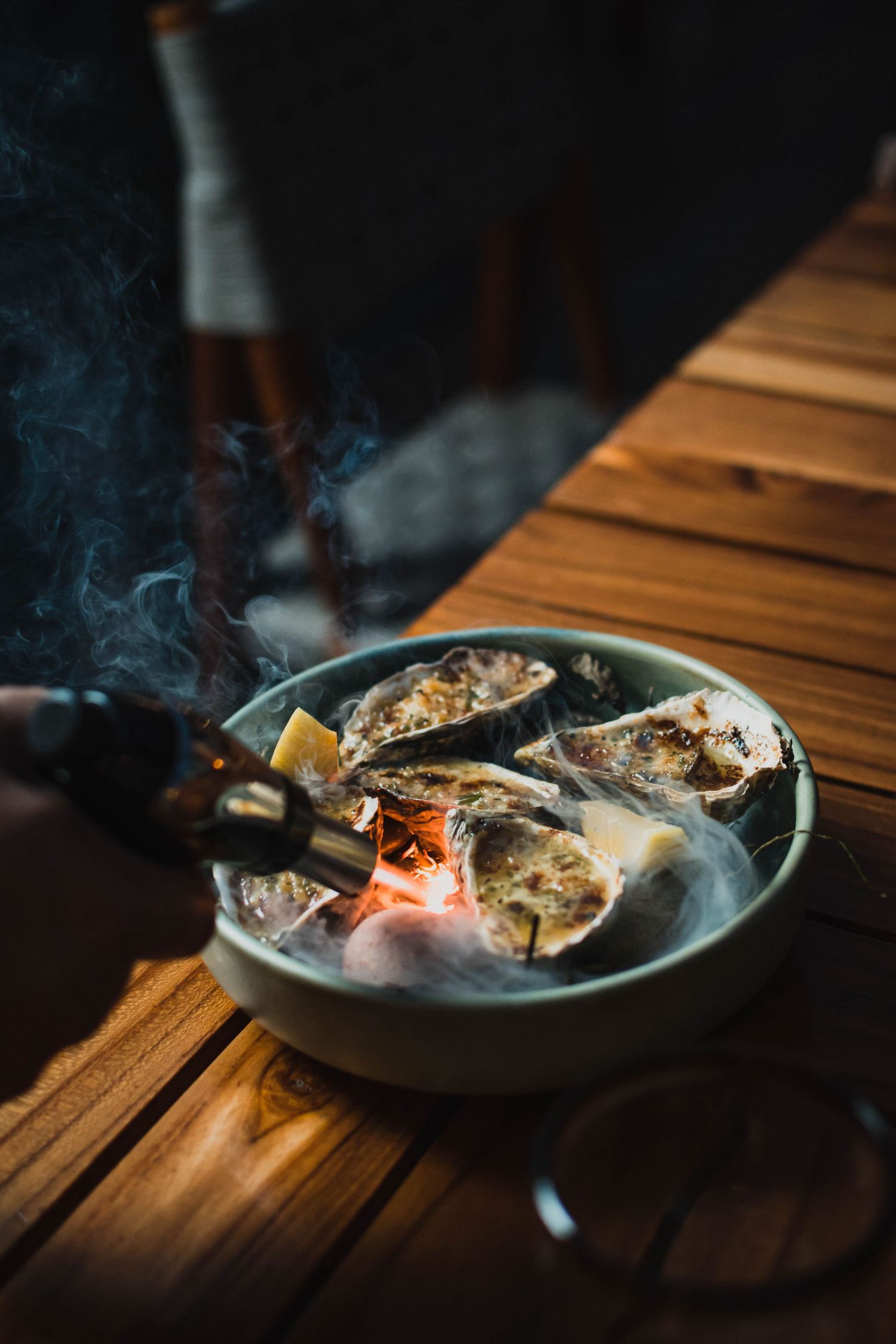 Smoked oysters on a dark wooden table with steam rising in moody lighting.