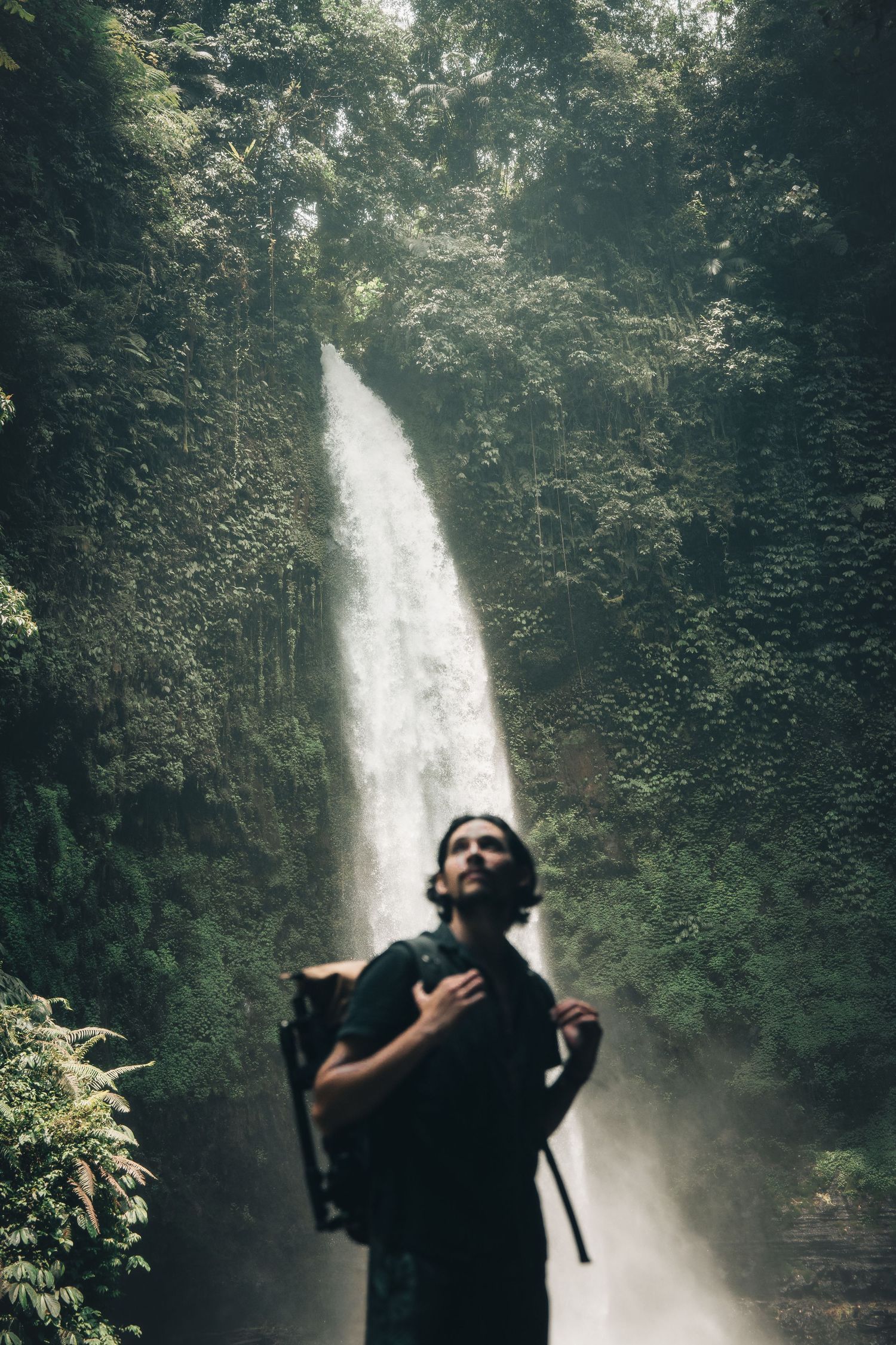 Silhouette of person photographing a towering waterfall in dense jungle setting.