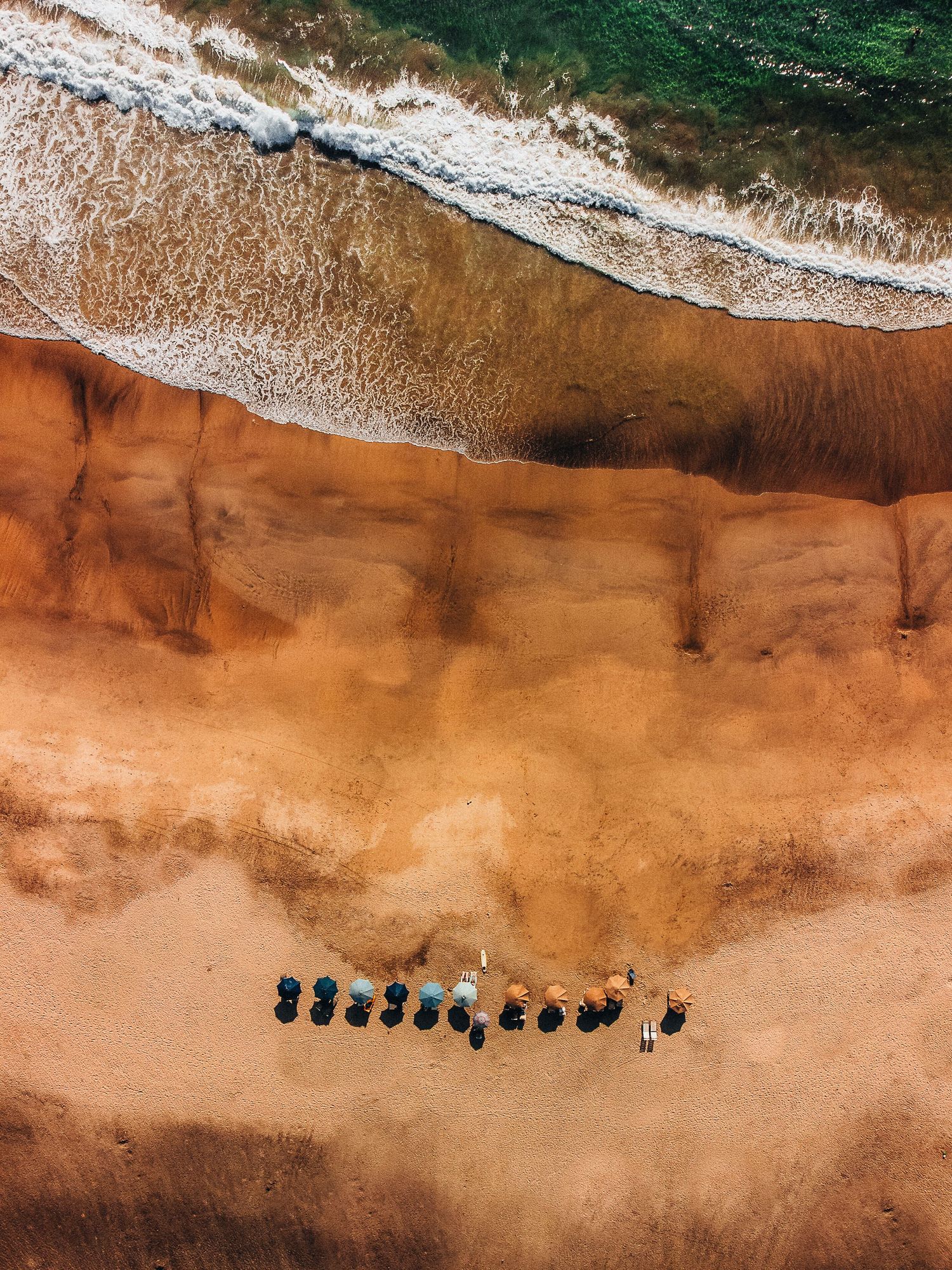 Aerial beach view showing orange sand meeting turquoise waters with people walking along the shore.
