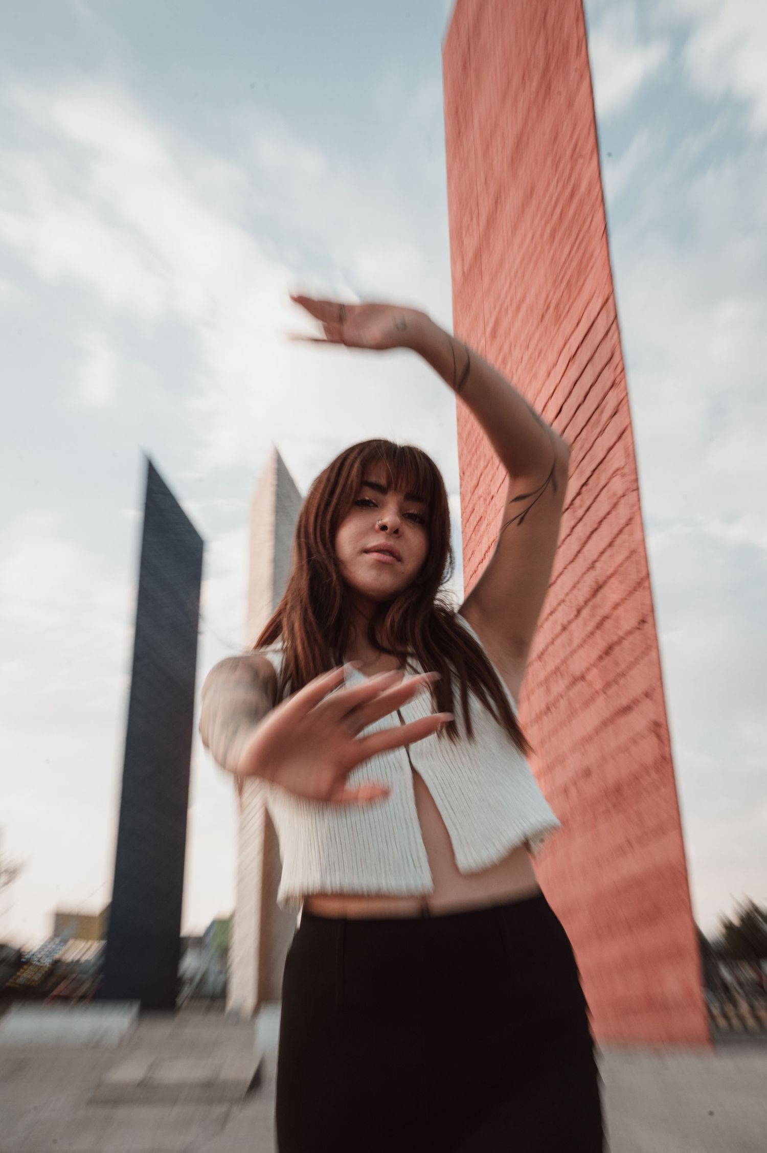 Artistic dance-like pose against geometric architecture with pink wall and cloudy sky.